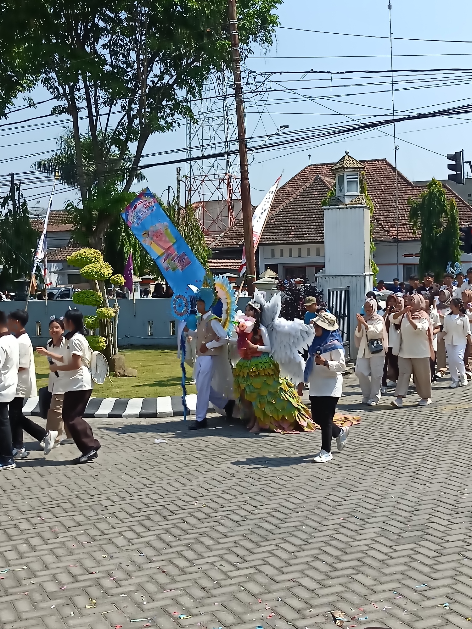 In Kediri City, Indonesia, a vibrant parade is underway on a paved street lined with trees and buildings. The event features participants in elaborate costumes, including a person dressed as an angel with large white wings and a flowing, multi-colored skirt. Several individuals in uniform, likely students, walk alongside the decorated participant, some holding banners, one of which displays "Pop Ice Colour Fest." The crowd watches the procession, with some individuals in the background also appearing to be in uniform or participating in the parade. The weather is sunny, with a clear blue sky overhead. The scene suggests a festive community event or school celebration taking place in an urban setting.