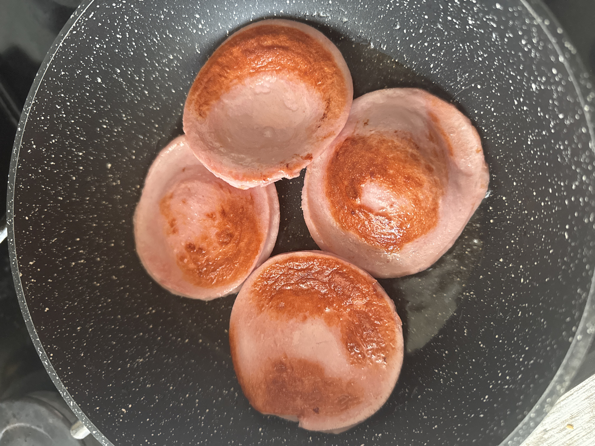 A top-down close-up shot reveals four slices of sausage cooking in a speckled frying pan. The sausages are lightly browned on one side and have a pink hue. The location is not discernible from the image alone.