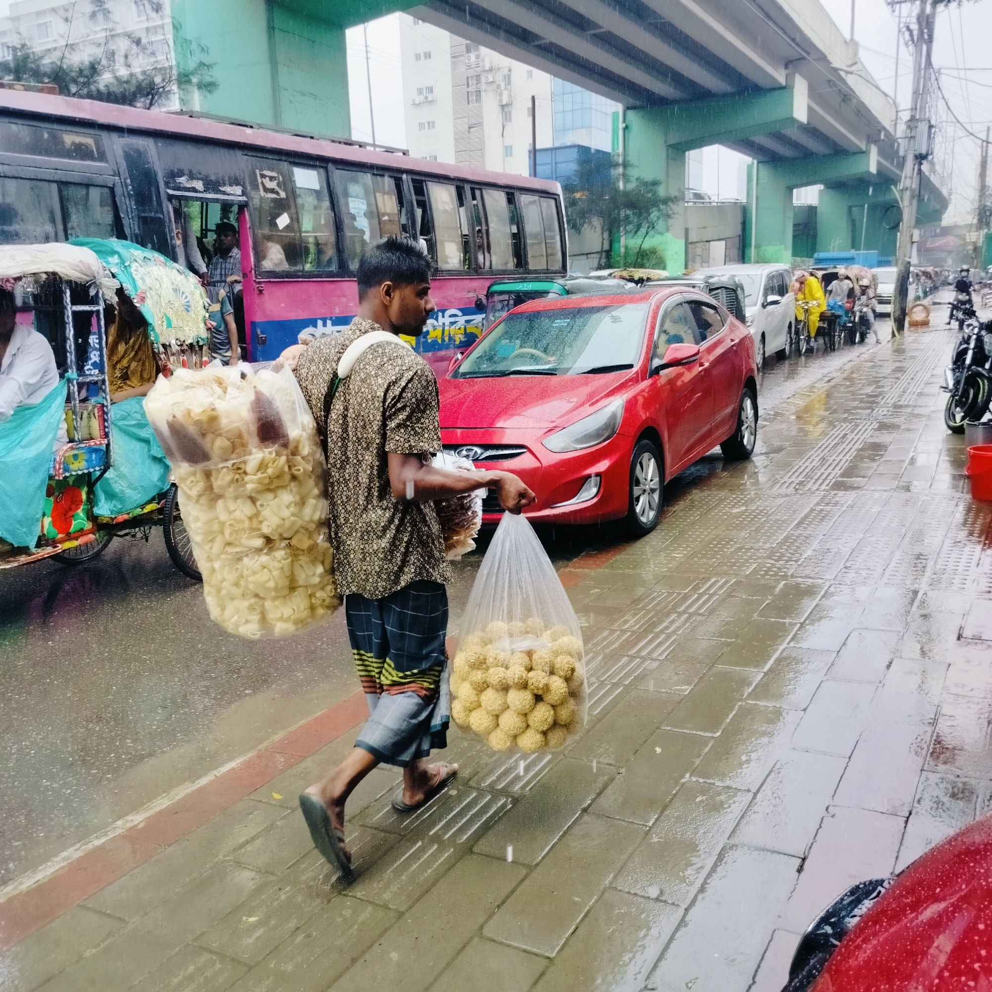 The image captures a street scene in Dhaka, Bangladesh, during rainfall. A man wearing a patterned shirt and checkered shorts is walking barefoot, carrying two large plastic bags of food items. The bag in his right hand contains round, yellowish items, possibly sweets or snacks, while the bag in his left hand holds lighter-colored, puffed snacks. He is walking on a wet sidewalk made of interlocking bricks, which reflects the surrounding environment.
In the background, a red car is parked on the wet road. Further behind, a pink and blue bus is visible, with passengers visible through its windows and an open door. The street is lined with various vehicles, including a bicycle rickshaw with passengers and several motorcycles. An elevated structure, likely part of a flyover, with green supports, runs overhead. The overall environment suggests a busy urban setting with a steady rainfall causing wet surfaces and reflections.