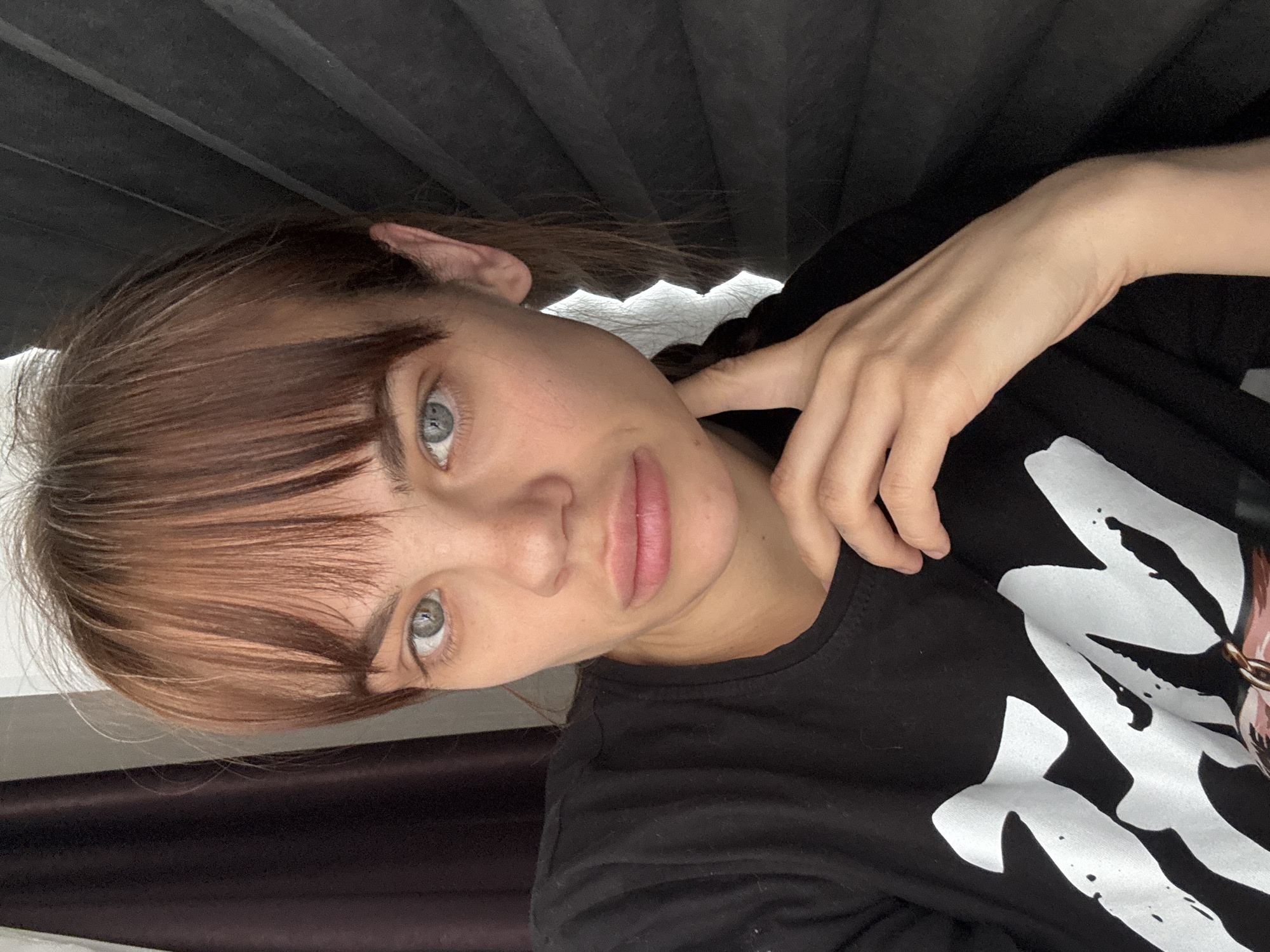 A young woman with light blue-grey eyes and light brown hair with bangs is depicted in an indoor setting. The image is framed as a close-up, focusing on her face and upper body. Her expression is neutral and calm. She is wearing a black crew-neck t-shirt that features a prominent white graphic design on the front, which appears to be a stylized symbol or abstract pattern. Her right hand (from her perspective) is gently placed near her neck or shoulder area. The background behind her consists of dark, pleated material, likely blinds or curtains, with a lighter, indistinct area visible to the upper left. The lighting is soft and even, suggesting it is either natural light from a window or diffused artificial lighting. The photograph is taken at an angle, giving the impression that the woman is lying down or tilting her head significantly. No legible text in English is visible on her clothing or in the background.