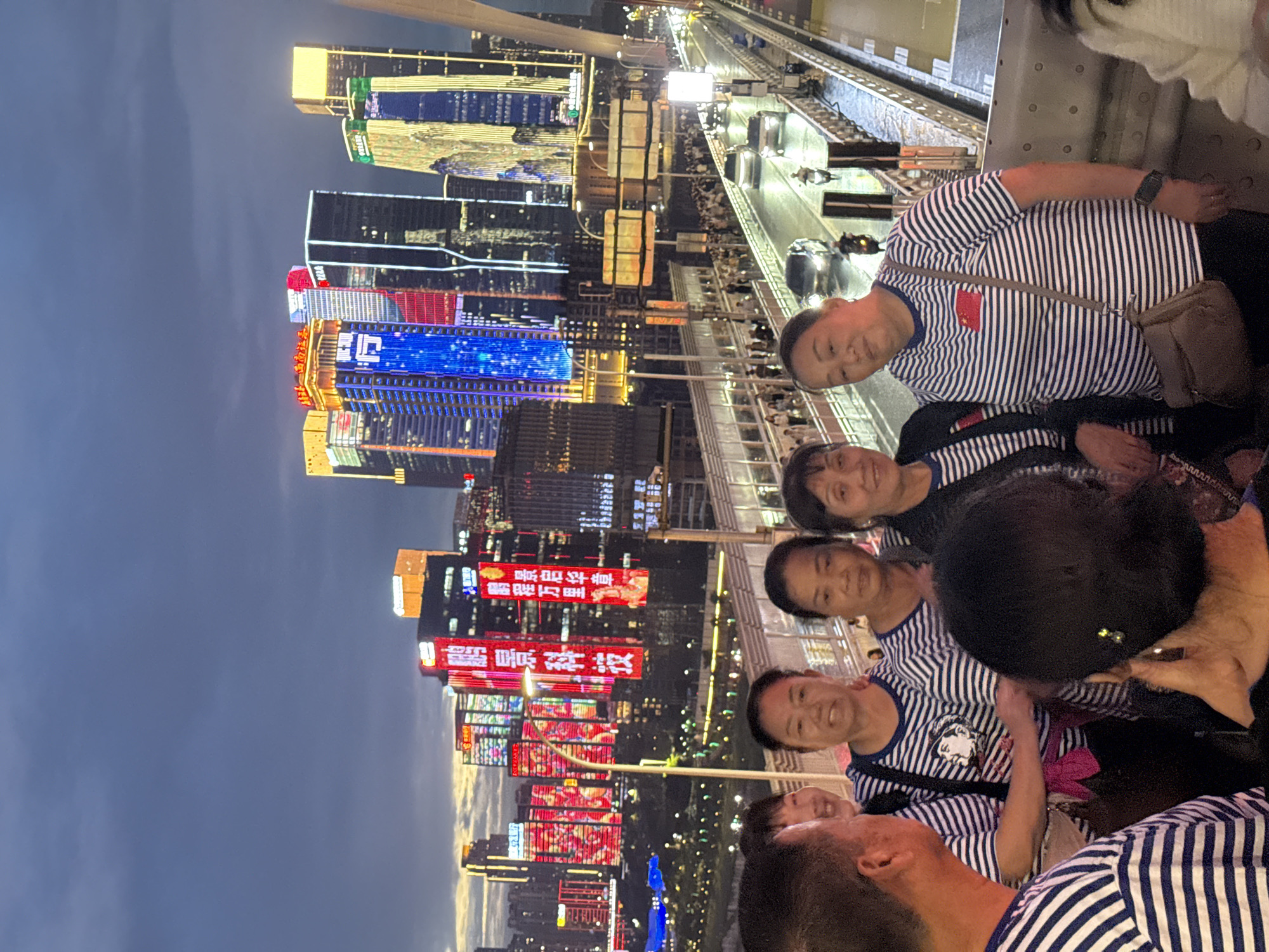 A group of six people stand on a bridge at dusk with a city skyline of illuminated skyscrapers in the background. The individuals are dressed casually, and several are wearing matching striped t-shirts. The skyline features numerous tall buildings with vibrant neon lights, including Chinese characters and abstract patterns. The bridge appears to be a busy thoroughfare, with cars and motorcycles visible on the roadway below. The sky is a dusky blue with hints of clouds. The location is identified as the Yuzhong District in China.