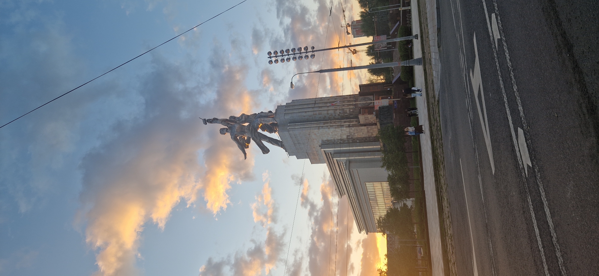 The image shows a wide shot of the "Worker and Kolkhoz Woman" monument in Moscow, Russia, at sunset. The sky is filled with dramatic clouds, illuminated by the setting sun with hues of orange and yellow. The iconic sculpture of a man and woman holding a hammer and sickle aloft stands proudly on top of a stepped, granite-clad pedestal, which is part of a larger building complex. To the left of the monument, a modern building with reflective windows is visible, contrasting with the classical design of the pedestal. In the foreground, a street with road markings and an asphalt surface is present. Several people are walking on the sidewalk in front of the monument and the building. A streetlamp with multiple lights and a regular streetlamp are also visible. In the background, trees and some construction cranes can be seen. The overall scene captures a blend of monumental Soviet-era art and architecture with the urban landscape of Moscow under a colorful sky.