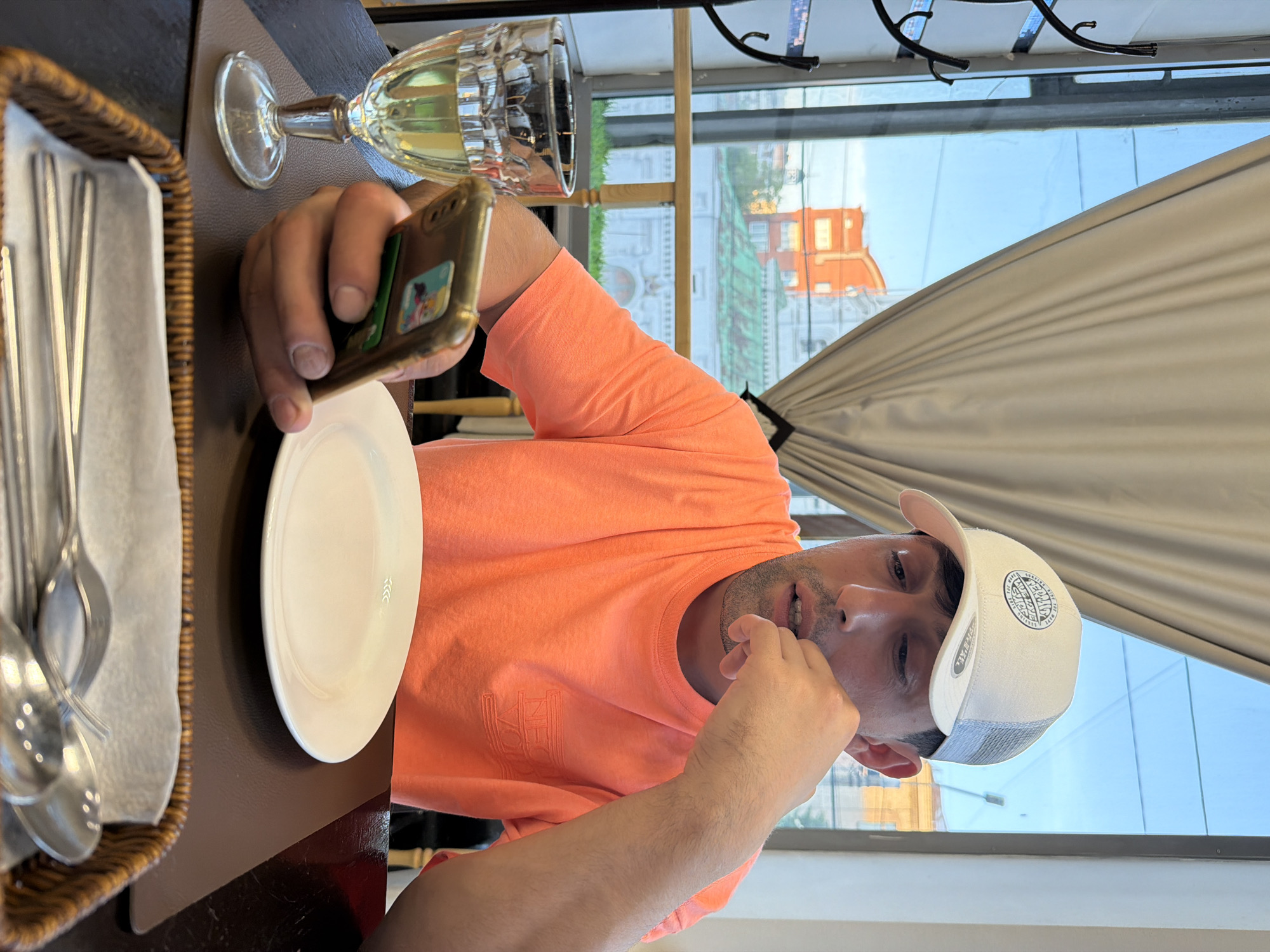 A man, appearing to be in his 20s or 30s, is seated at a dark brown table in what seems to be a restaurant or cafe, possibly in Krasnoyarsk, Russia. He is wearing a bright orange short-sleeved t-shirt, which has a stylized, partially visible logo on the left chest area, and a white baseball cap with a mesh back. The cap features a circular black logo with text including "Rippin' & Skippin'" and "Est. MMIV," while "TYR & ALL" is visible on its side.
The man is holding a gold-colored smartphone in his left hand, with a small card or item tucked into its case, and appears focused on the device. His right hand is raised near his mouth, suggesting a thoughtful or casual gesture. In front of him on the table is an empty white plate. To his left, a wicker basket holds several spoons and forks on a white napkin. A stemmed glass, partially filled with a pale yellow beverage (likely white wine), is situated near his left hand.
Behind the man, a large window reveals a bright blue sky and reflections of urban buildings, some with distinctive red or orange roofs. A light-colored curtain is drawn across part of the window. The clear sky and bright reflections suggest it is daytime, likely afternoon or early evening. The scene conveys a casual dining environment.