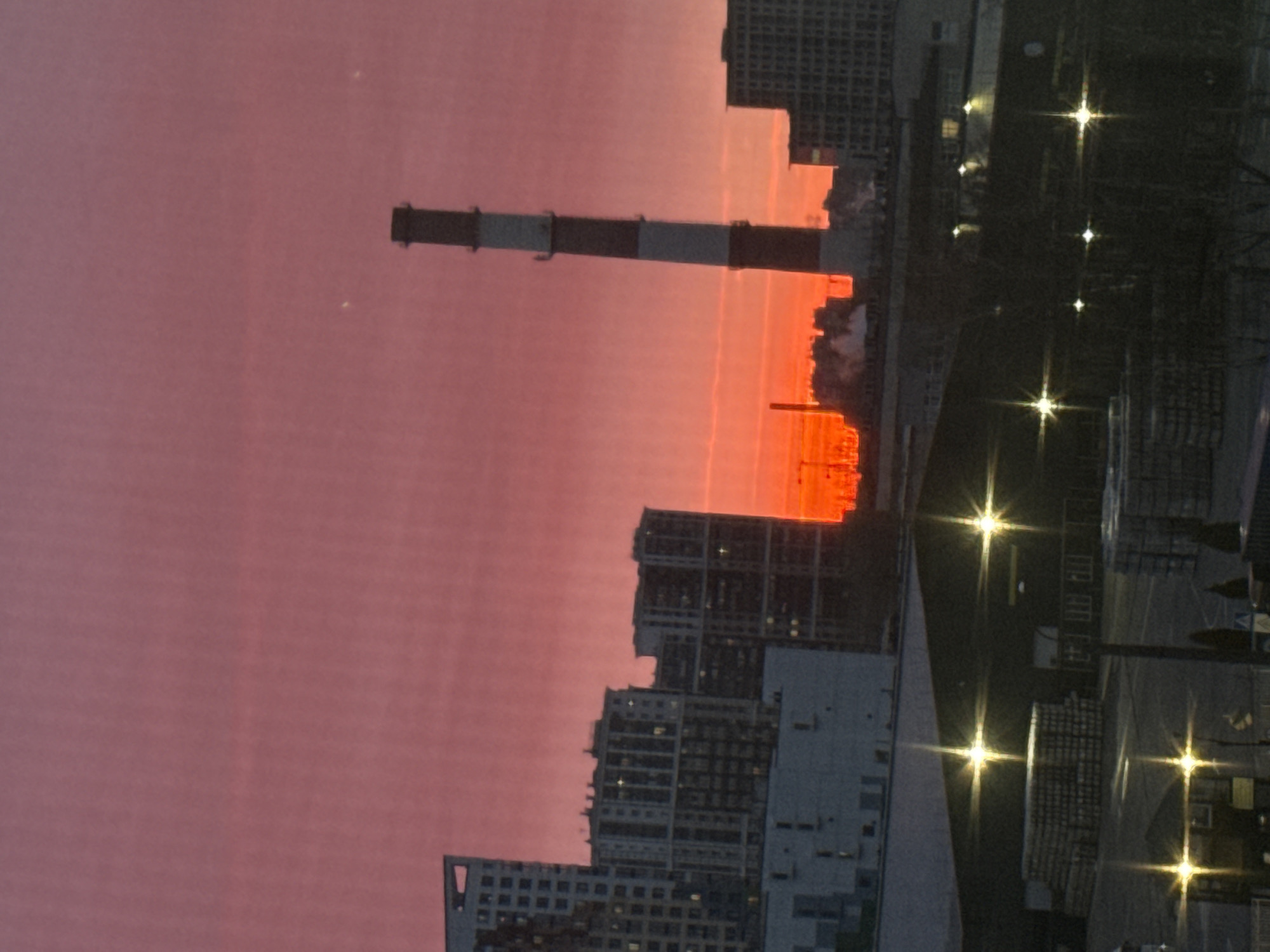 The image shows a cityscape at sunset with a prominent smokestack extending vertically. The sky is a gradient of pink and orange. Buildings of various sizes are visible, some partially obscured by the setting sun. Lights are visible from streetlights. The location is Saint Petersburg, Russia.