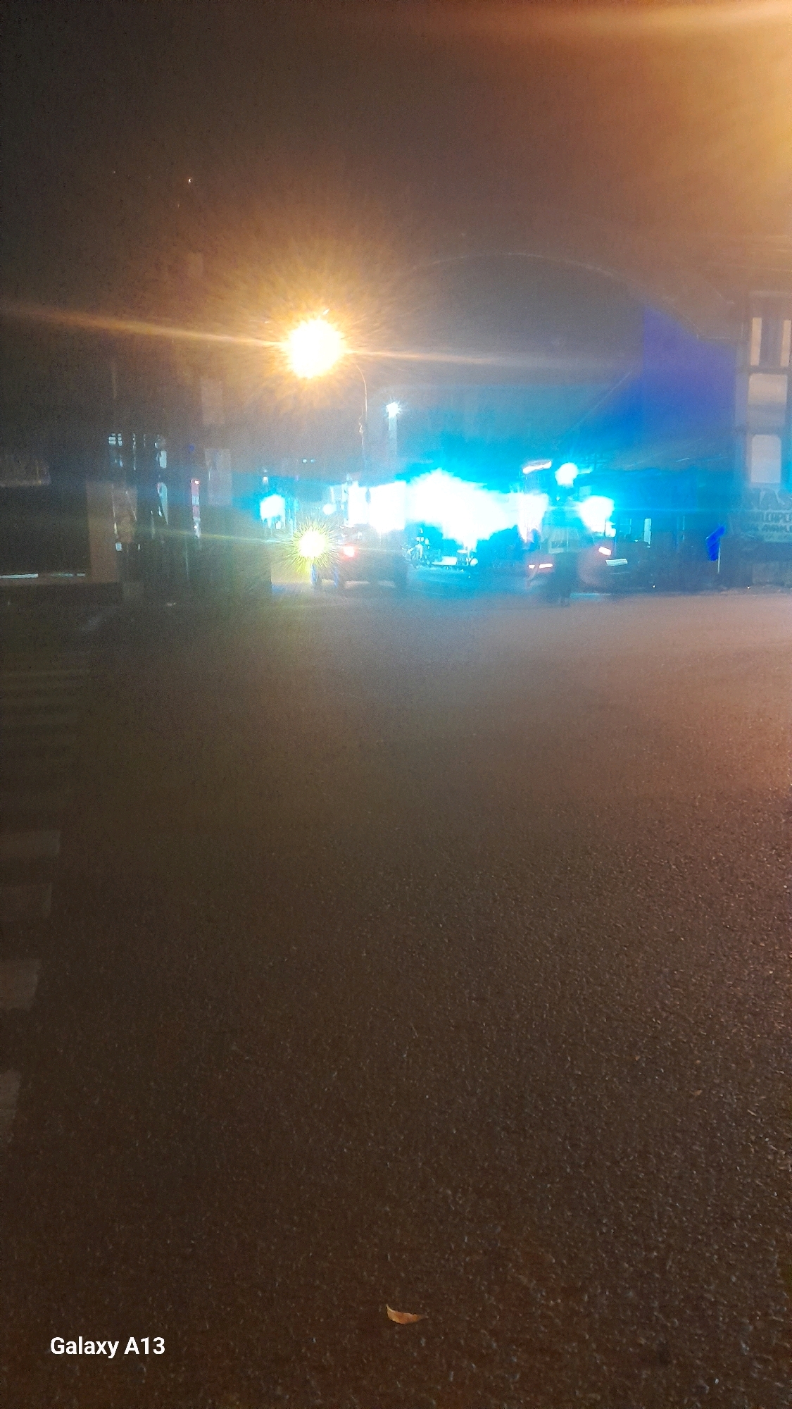 The image depicts a street scene at night in Ngembalrejo, Indonesia, characterized by bright artificial lighting. A prominent streetlamp with a warm, intense glow is visible on the left side, casting significant light and lens flare across the upper portion of the frame. Below and to the right, a cluster of bright blue and white lights emanates from what appears to be a vehicle or group of vehicles, possibly indicating emergency services or a traffic incident. The road surface, wet and reflective, shows faint zebra crossings on the left. A single fallen leaf rests on the asphalt. The background is obscured by darkness and the intense lights, but suggests the presence of buildings or structures. The overall impression is of a well-lit, possibly busy, urban street at night.
