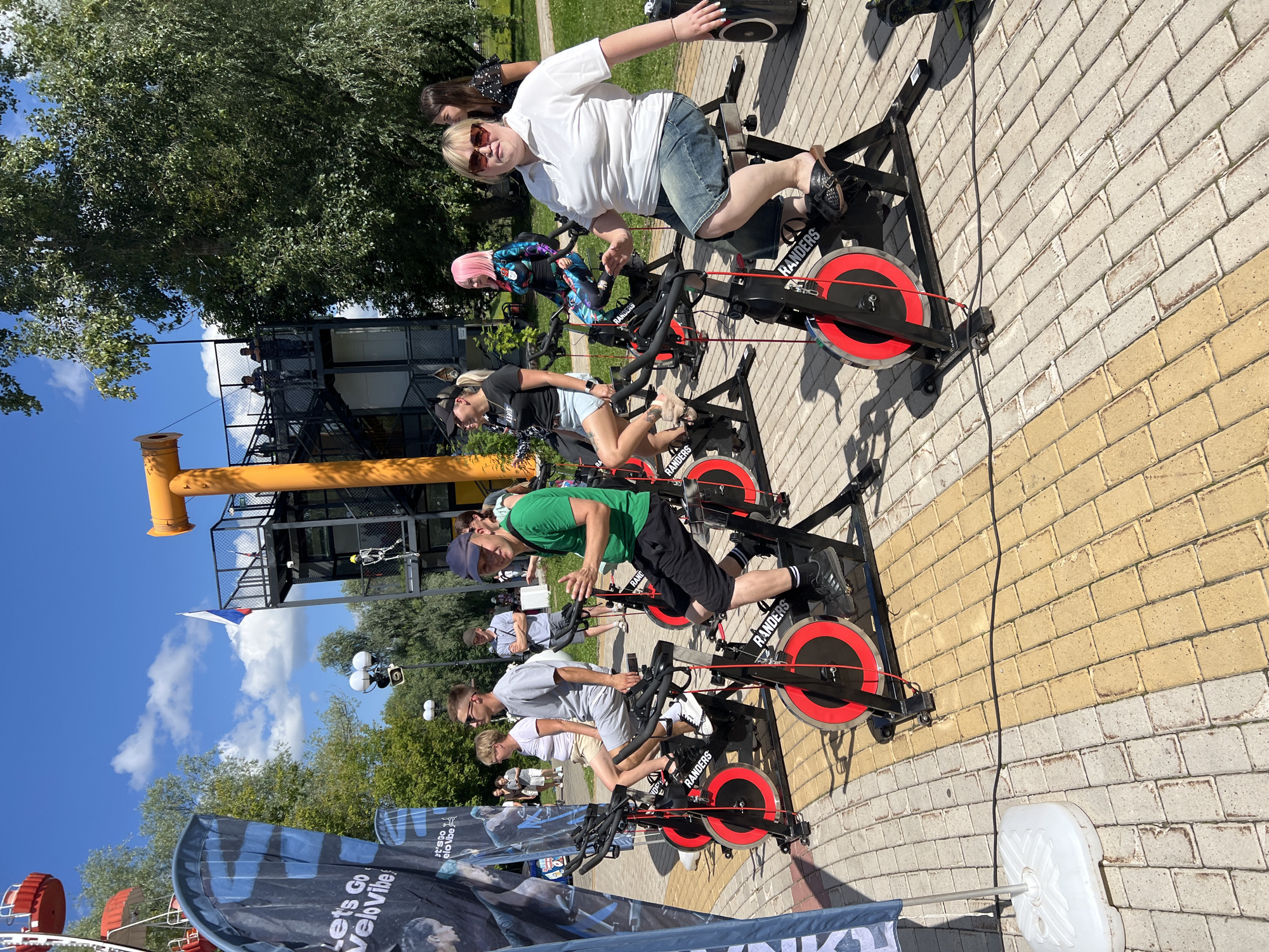 In Cheboksary, Russia, on a sunny day with a partly cloudy sky, a group of people are engaged in an outdoor stationary cycling event. Several individuals are seated on black exercise bikes with bright red flywheels, actively pedaling. The bikes have the branding "RANDERS" visible. The ground is paved with a mix of light beige and yellow rectangular bricks, creating a patterned walkway.
In the background, a tall yellow structure, possibly part of an amusement park or recreational facility, stands prominently against the sky. To its left, a Ferris wheel, with red gondolas, is partially visible. A Russian flag flutters from a pole near the yellow structure. Trees with lush green foliage surround the area, providing some shade.
The participants are dressed in casual summer attire. One woman in the foreground wears a white polo shirt and denim shorts, while another person in the foreground is wearing a green t-shirt and black shorts. Other individuals are also visible on bikes, some with distinctive hairstyles like bright pink hair. A large blue banner with the text "Let's Go VeloVibe" is partially visible on the left side of the image. The overall atmosphere suggests a public event or fitness activity taking place outdoors in a park-like setting.
