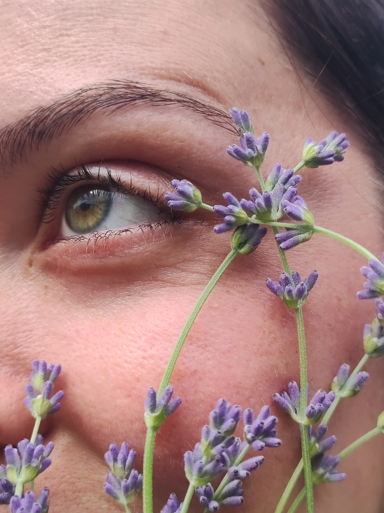 The image is a close-up, outdoor shot of a person's face, with a focus on their eye and cheek, partially obscured by sprigs of lavender. The person's left eye, with green and yellow irises and dark lashes, is looking to the right. The skin is lightly textured, with some pores visible. The lavender flowers are small and purple, with green stems. The background is a blurred expanse of the person's skin and dark hair. No specific location in the city of Фоки, Russia, can be determined from the image.