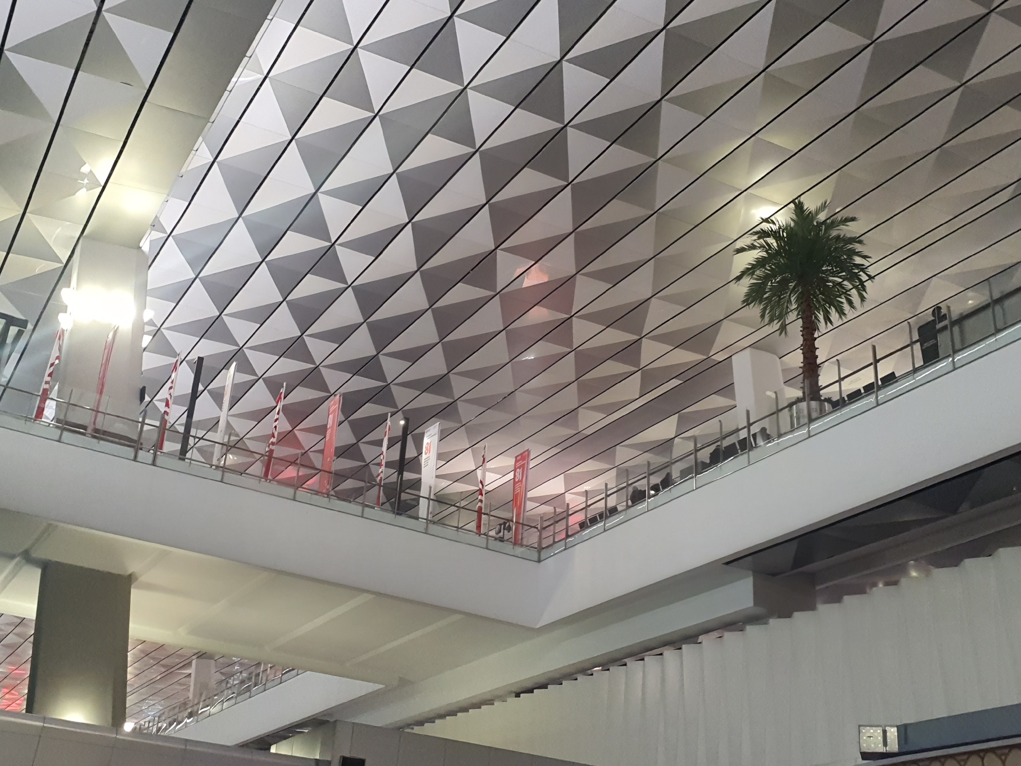 The image depicts the interior of a modern building with a distinctive triangular-patterned ceiling. The ceiling is lit from above, casting subtle shadows and highlights on the geometric surfaces. In the midground, a multi-level structure is visible, with balconies or walkways extending across. Several flags, predominantly red and white, are displayed on the upper level, suggesting a national or event-related context.
A lone palm tree stands on the right side of the image, its fronds reaching towards the ceiling, adding a touch of nature to the man-made environment. On the upper walkway, several figures can be seen, some standing and others possibly seated, their silhouettes indistinct against the lighting. One individual is clearly visible standing near the palm tree, looking towards the right. The overall impression is of a spacious, architecturally interesting interior, possibly an airport terminal or convention center.
This scene is located in the city of Tangerang, Indonesia, specifically within the Terminal 3 area of Soekarno-Hatta International Airport.
