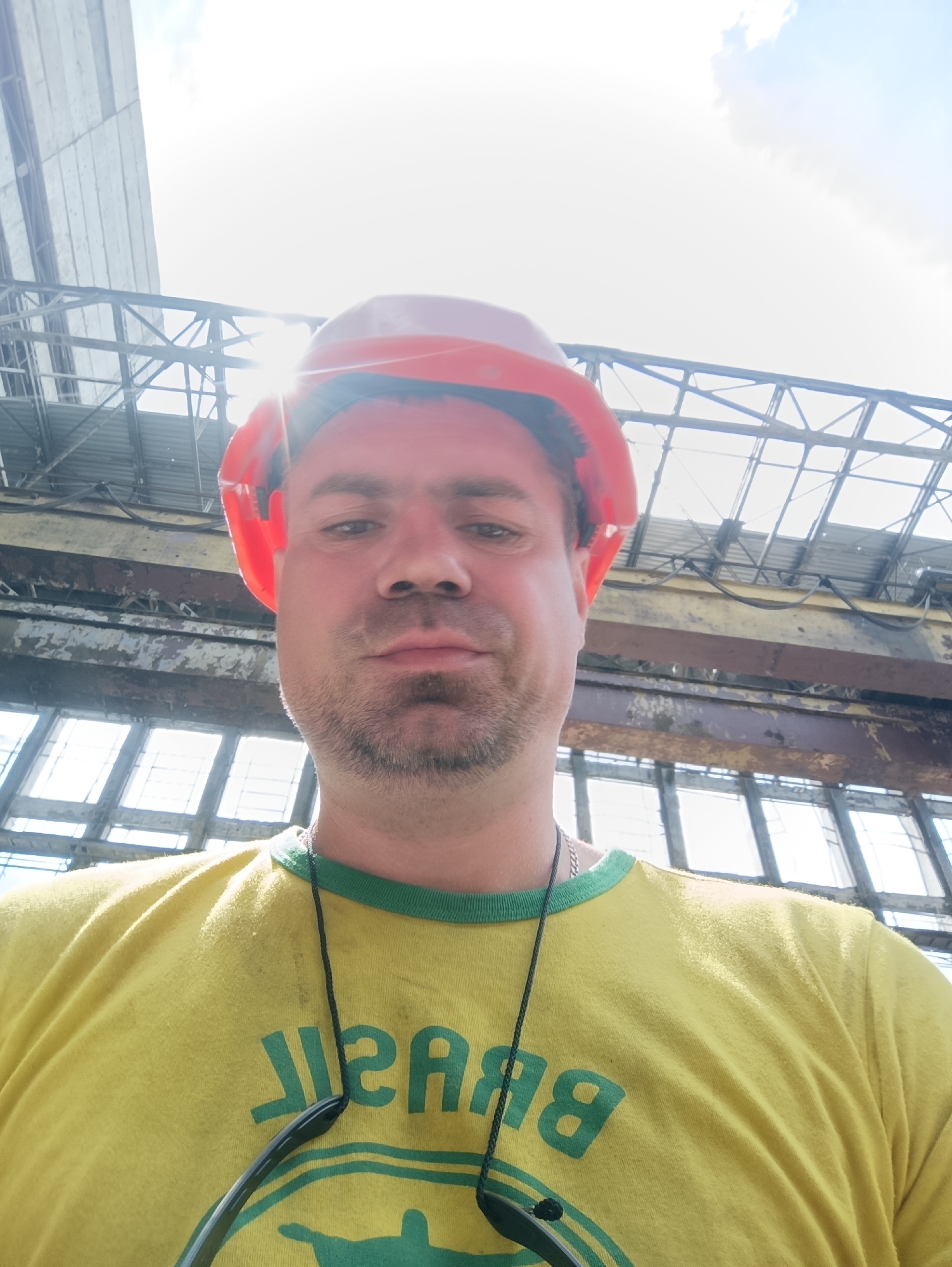 A man wearing a bright orange hard hat and a yellow t-shirt with green lettering and trim is standing in front of large industrial windows. The t-shirt has green text that appears to be "BRASIL" printed on it. The man has short brown hair and a beard. He is wearing a necklace. Sunglasses hang from the neckline of his shirt. The background consists of a large, industrial structure with exposed metal beams and corrugated roofing. Large, multi-pane windows are visible behind him. The sky above is bright and partially cloudy, with sunlight streaming through the roof structure, creating lens flare in the image. The overall scene suggests an indoor industrial or construction environment.