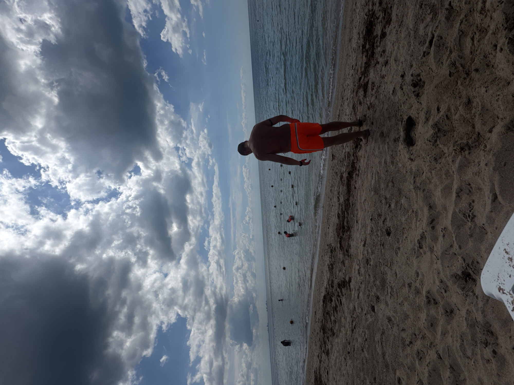 The image depicts a man standing on a sandy beach in Yevpatoriya, Ukraine, near the sea. He is wearing orange swim trunks and is facing the water. The sea is calm, with some buoys visible in the water. The sky is partially cloudy with dark rainclouds to the left. The image is shot from a high angle. The sand is dark and coarse. Part of a white object is visible in the right corner.