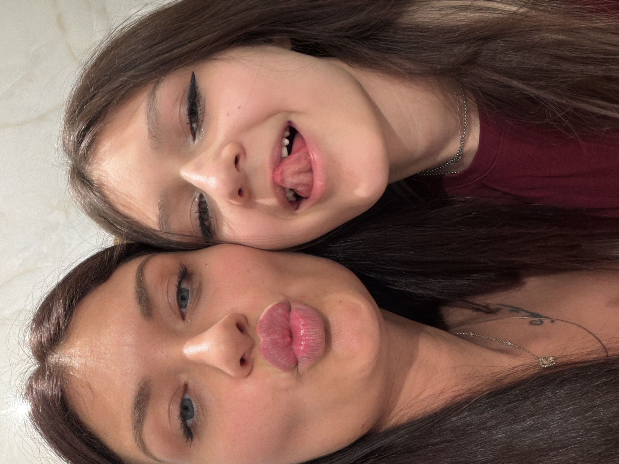 A close-up, vertically oriented selfie features two females with dark hair, positioned side-by-side. The person in the upper half of the frame, appearing younger, has dark hair and visible eyeliner, and is making a playful, silly face with her tongue sticking out. She wears a dark red top and a thin silver chain necklace. Her expression is one of amusement.
The person in the lower half of the frame, appearing older, has dark hair and blue eyes, and is making a "puckered lips" or "kissing" face. Her lips appear glossy. She wears a thin silver necklace with a square, possibly diamante, pendant. A partial tattoo is visible on her neck or upper shoulder, partly obscured by her hair.
The individuals are captured in a tight crop, showing their faces side-by-side, suggesting an intimate or familial connection. The background is a light-colored, out-of-focus surface, possibly an indoor wall or ceiling. The lighting is soft and even. No text is visible.