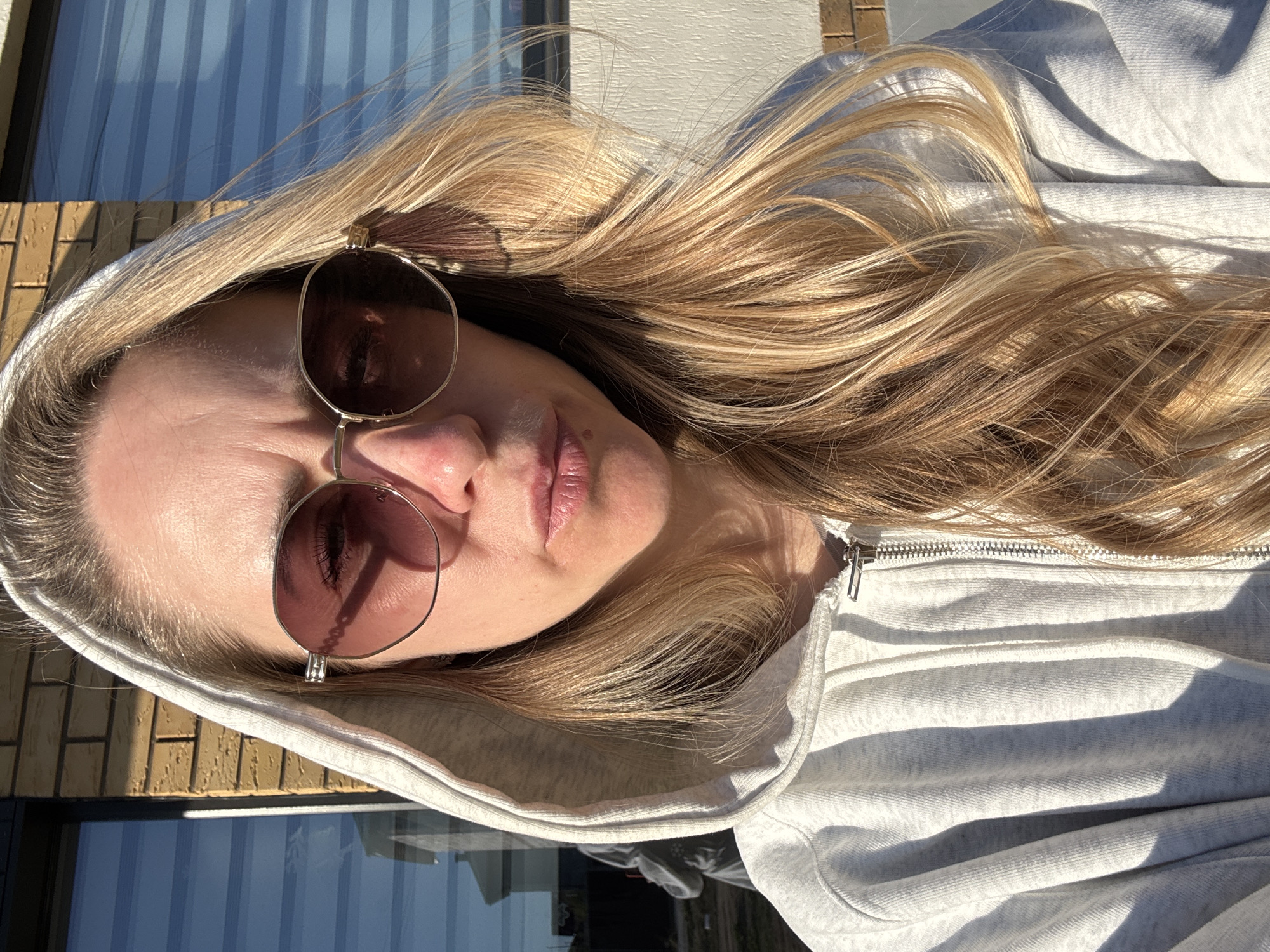 A woman with blonde hair and sunglasses is wearing a hooded sweatshirt. The background includes a building with windows. The photo was taken outside in sunny conditions. There is no verifiable evidence to support the location being Chaikovsky, Russia.