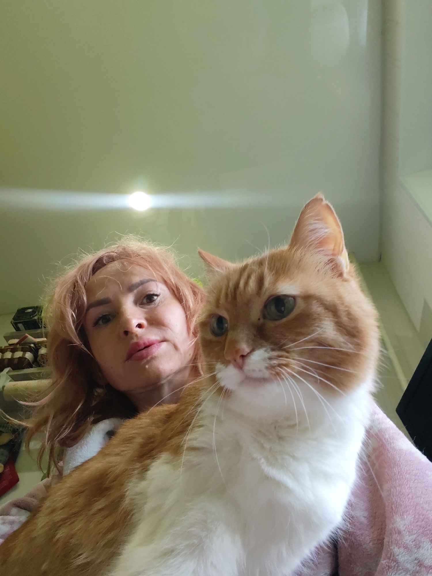 A low-angle, close-up selfie captures a woman and an orange and white cat. The woman, with shoulder-length, light pink hair, is positioned slightly to the left, looking towards the camera with a neutral expression and light makeup. Her mouth is slightly parted, revealing a light shimmer. The cat, mostly filling the right side of the frame, is an orange tabby with white chest and paws, its head tilted slightly to the left, and its eyes are a striking green. The background is indistinct, showing a pale green ceiling with a bright overhead light and some shelving with boxes on the left side, hinting at an indoor domestic setting. The woman appears to be wearing a pink and white fluffy robe or blanket. The image is taken from a perspective that suggests the woman is holding the cat. No specific location in Chaikovsky, Russia is identifiable from the visual cues in the image.