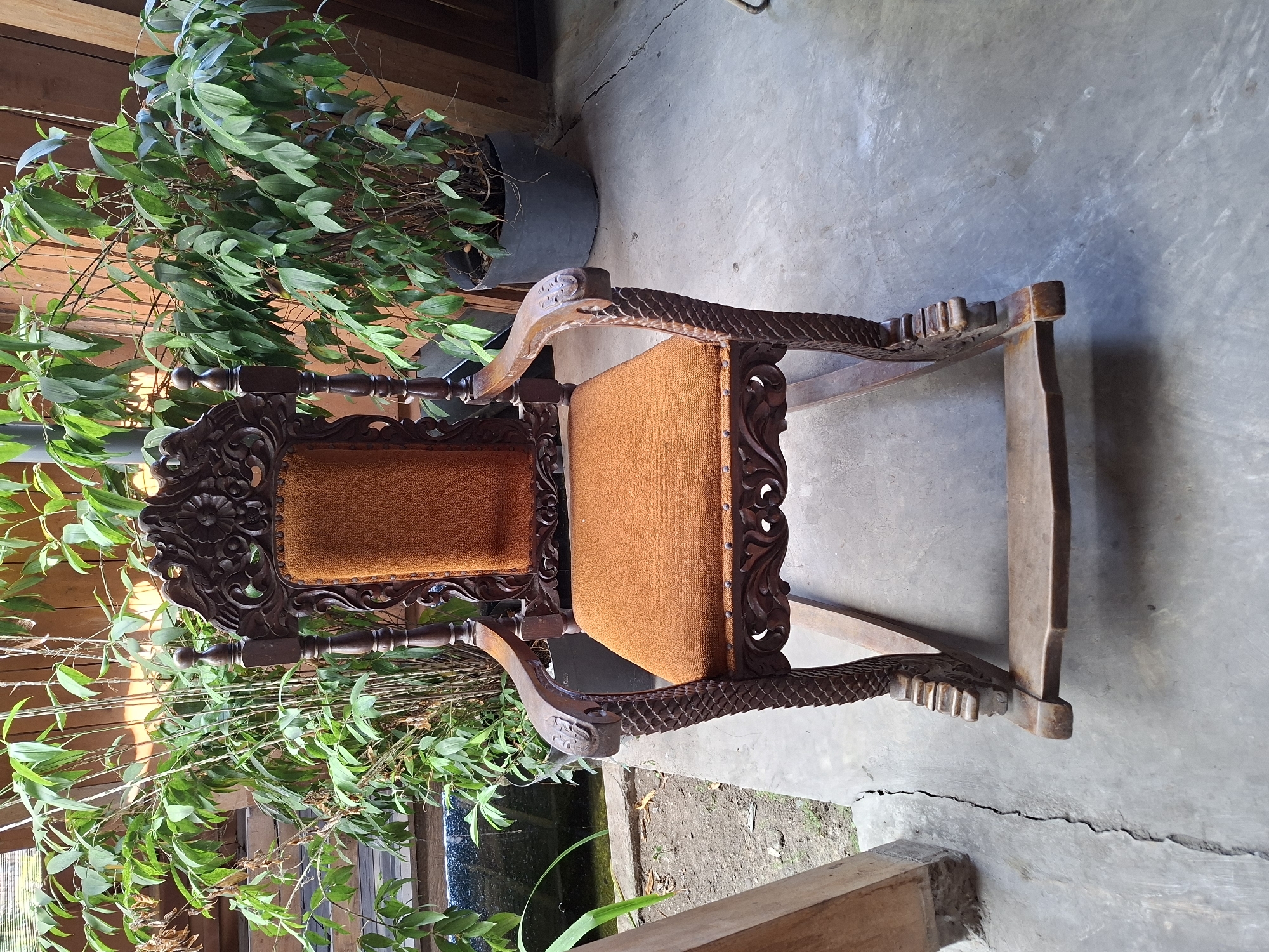 This image shows a carved wooden rocking chair with an orange upholstered seat and back, positioned outdoors on a concrete patio. Behind the chair, there is lush green foliage, including what appears to be bamboo and potted plants. A wooden fence or structure is visible in the background. The scene is lit by natural light, suggesting it is daytime. The setting appears to be a garden or courtyard area, possibly in a residential or commercial property in Purworejo, Indonesia. No people are present in the image.