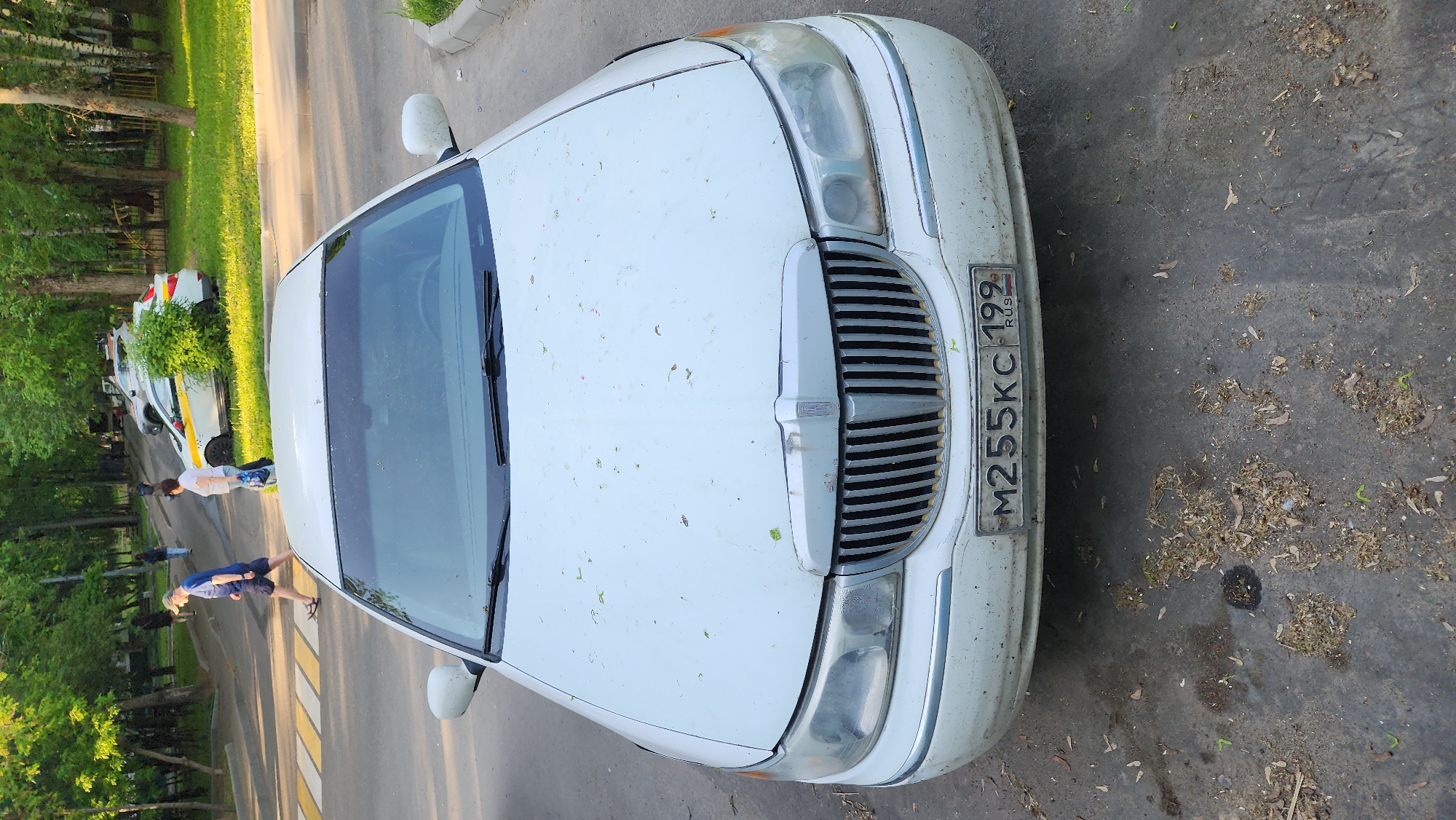 A white Lincoln Town Car with the Russian license plate "M255KC 199 RUS" is parked on a paved surface covered with scattered debris, possibly in Moscow, Russia. The car appears to be dirty, with leaves and other debris covering its hood. Two people are walking across a crosswalk in the background near a white van. Trees and green foliage are visible on both sides of the crosswalk.