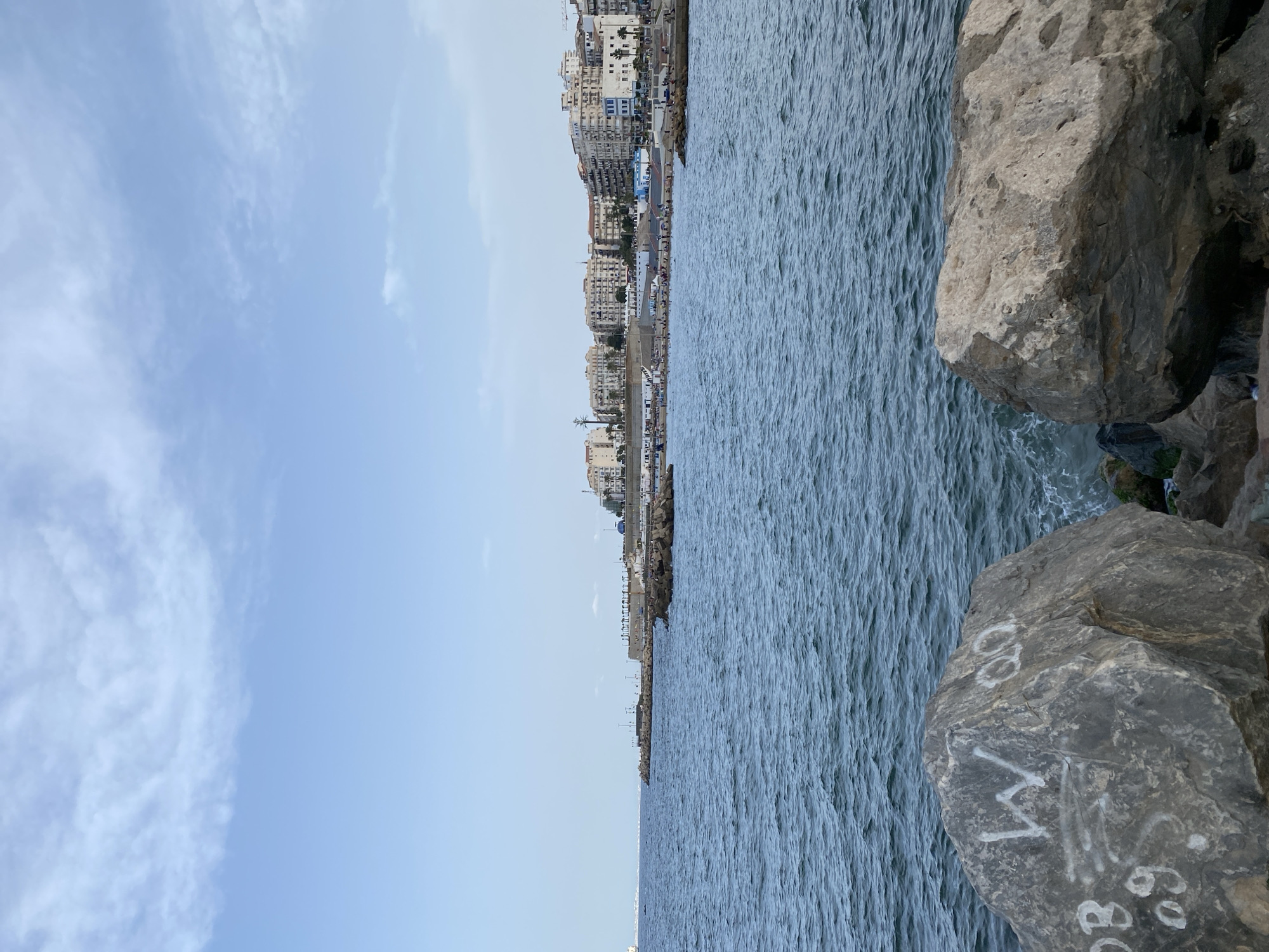 The image shows a view of the sea from a rocky shoreline in Beau Fraisier, Algeria. In the foreground, large gray rocks with graffiti are visible. The sea is a deep blue with ripples. In the background, a coastline with buildings and palm trees can be seen, with people visible on the beach and along the promenade. A ship is in the distance on the left side of the frame. The sky is blue with scattered white clouds.