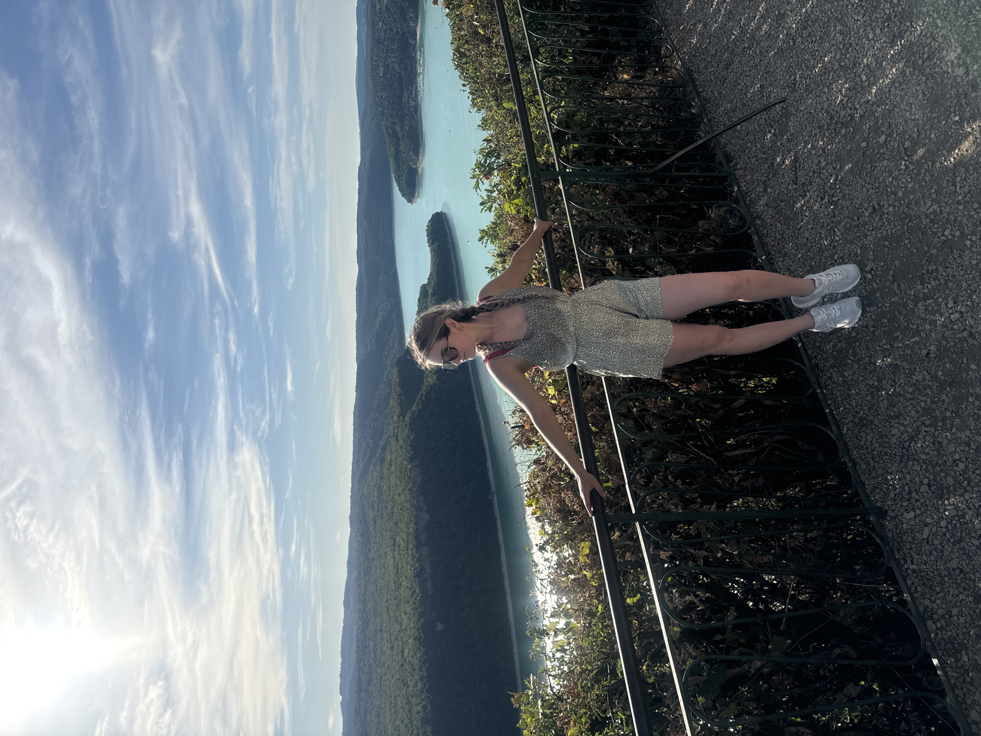 A young Caucasian woman with blonde hair in two braids, wearing a patterned romper and white sneakers, stands with her arms resting on a black metal railing. She is looking off to her left, with a scenic backdrop of a turquoise lake, lush green forested hills, and a blue sky with wispy clouds. The foreground shows a gravel path. The location is the Belvedere de La Forge in Moirans-en-Montagne, France, offering a panoramic view of the Lac de Vouglans.