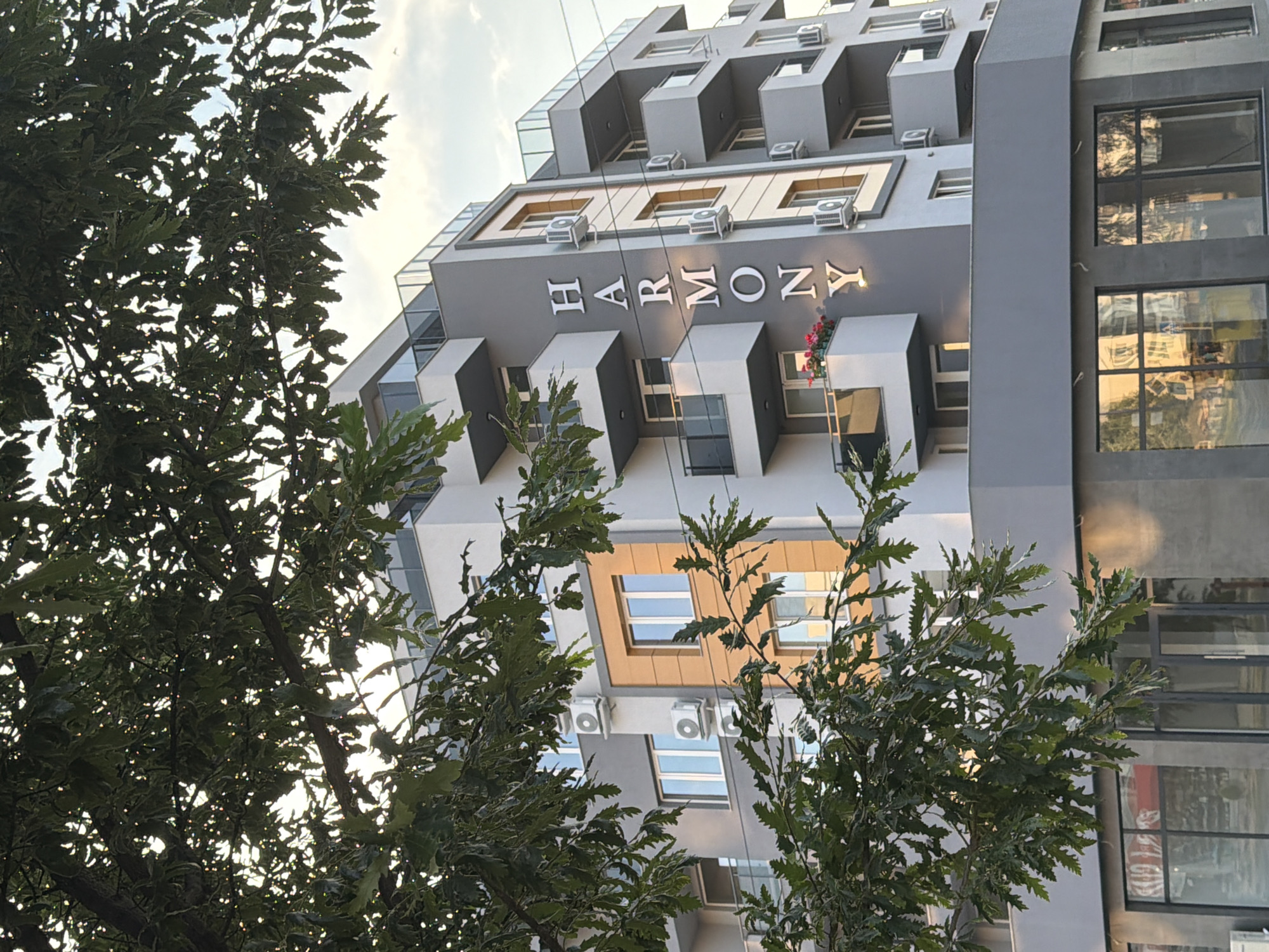 A modern residential building, identified as "HARMONY," is depicted in Pazardzhik, Bulgaria. The building features a contemporary design with multiple balconies, gray and orange facades, and numerous air conditioning units. The foreground is partially obscured by the branches and leaves of a large tree, suggesting an outdoor setting on a street or public space. The sky above is partly cloudy, indicating daylight conditions. The ground floor of the building appears to house commercial establishments with large glass windows, reflecting the surrounding environment. There are no people visible in the image.