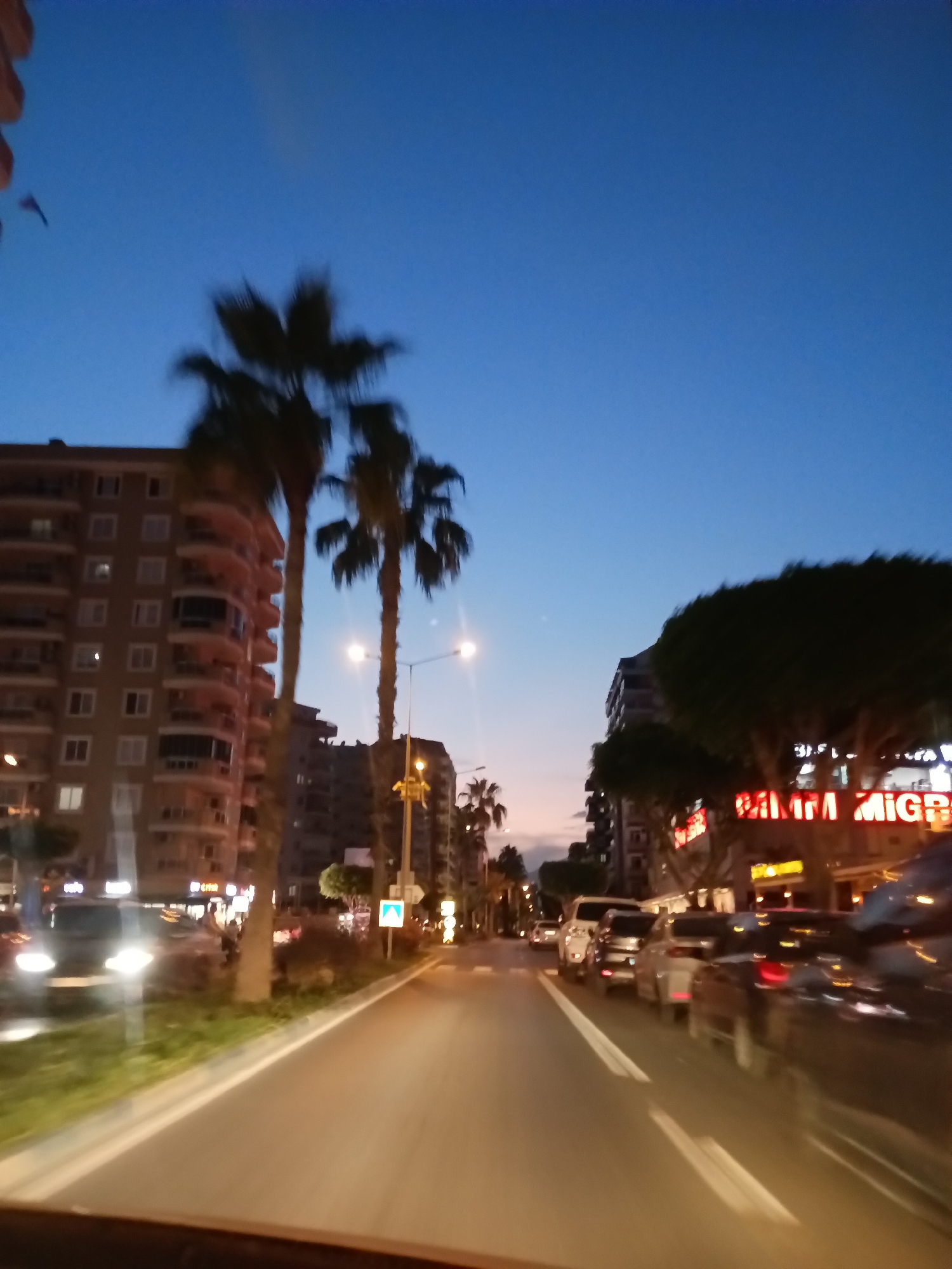 This image captures a street scene in Alanya, Turkey, at dusk. A multi-lane road with visible lane markings runs through the center of the frame. Cars are present on the road, suggesting moderate traffic. On the right side of the road, a brightly lit neon sign reads "BİM MARKET," indicating a commercial establishment. Palm trees are scattered throughout the scene, with their silhouettes visible against the darkening sky. Residential buildings line the left side of the street, characterized by multiple floors and balconies. Streetlights are illuminated, casting a warm glow on the surroundings. The sky transitions from a lighter hue near the horizon to a deep blue at the top of the frame, signifying the onset of night. The overall impression is a bustling urban environment in the twilight hours.