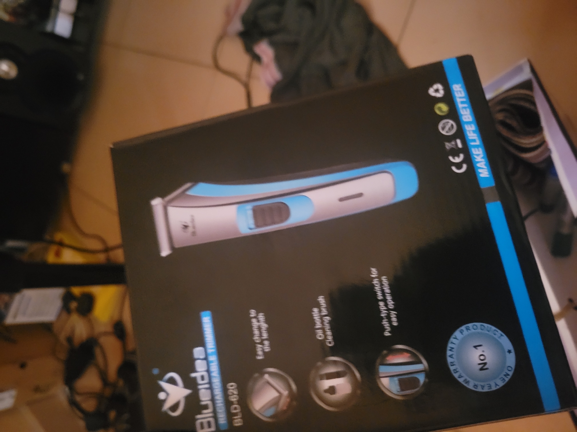 The image shows a close-up, slightly blurry view of a product box for a "Blueidea RECHARGEABLE TRIMMER BLD-620". The box is primarily black with blue accents. The front of the box displays the brand logo, the product name, and several circular icons with text indicating features such as "Easy change to the length", "Oil bottle Cleaning brush", and "Push-type switch for easy operation". There is also a circular "ONE YEAR WARRANTY PRODUCT No.1" badge. On the right side of the box, a light blue and silver electric trimmer is depicted. Towards the bottom right corner, there are several certification symbols and the text "MAKE LIFE BETTER". The background of the image is out of focus and appears to be an indoor setting with some electrical cords and fabric visible. The scene is static and there are no people or animals present. The lighting is moderate, with some reflections on the glossy surface of the box.