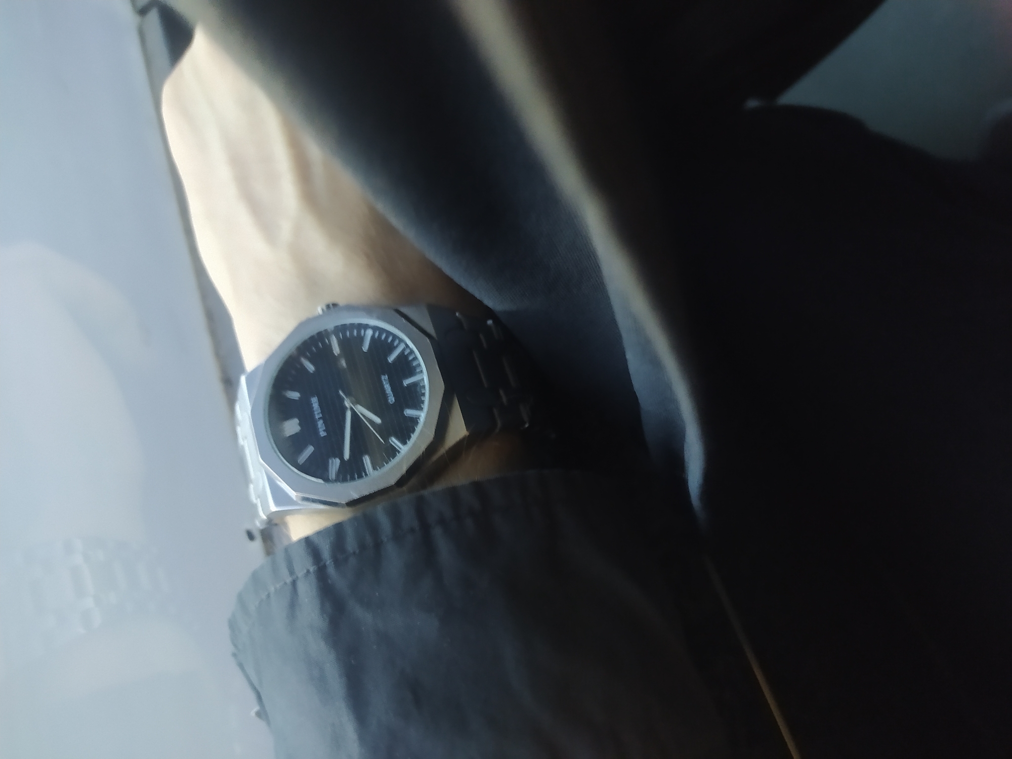 A close-up shot captures a silver metal wristwatch with a black dial on a person's wrist. The watch band is also made of silver metal. The watch face features white hour markers and hands, with "PIN TIME" and "QUARTZ" printed on it. The person's arm is covered by a dark gray or black garment, possibly a jacket. The background is blurred and appears to be a light-colored, reflective surface, possibly a window or a dashboard, with a faint reflection of what could be a car's interior. The scene is lit by natural light, creating a slightly hazy atmosphere. The location is unknown.