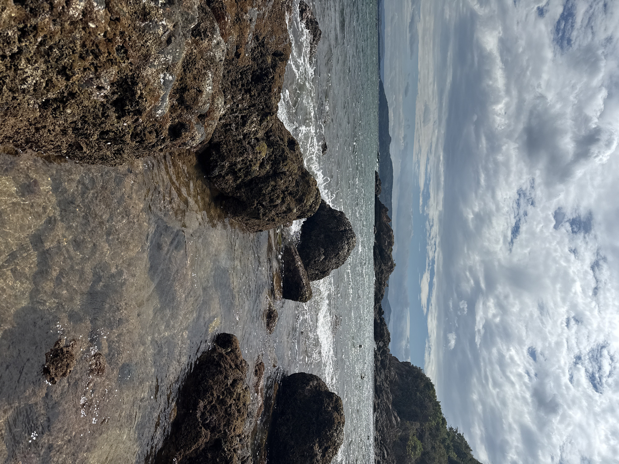 The image shows a rocky shoreline on a cloudy day. In the foreground, large, dark brown rocks are partially submerged in clear, shallow water. Some of these rocks are covered in a rough, textured growth, possibly algae or barnacles. Small waves are breaking against the rocks, creating white foam. The water is calm beyond the immediate shoreline, and a larger body of water stretches to the horizon. In the background, a rugged, tree-covered hillside rises from the water's edge. The sky is filled with dramatic, grey and white clouds, with patches of blue sky visible. The scene appears to be located in a natural coastal area. Based on the available context, this location is identified as Song Cau City, Vietnam. There are no people or specific man-made objects visible in the scene.