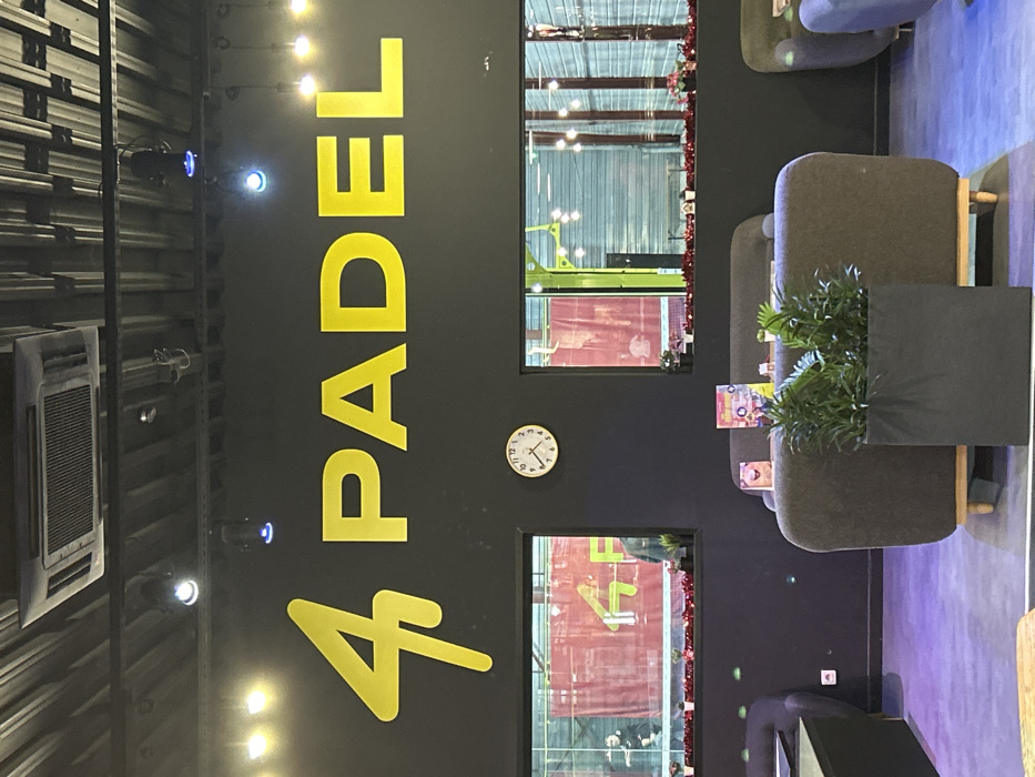 A brightly lit interior features the words "4 PADEL" in large yellow letters on a dark wall. Two windows showcase a padel court with players. A white clock is mounted on the wall. To the right, there are lounge chairs with small tables holding leaflets, along with a tall, dark planter with green plants. An air conditioning unit is visible on the left wall. The overall impression is a modern, stylish padel club reception area.