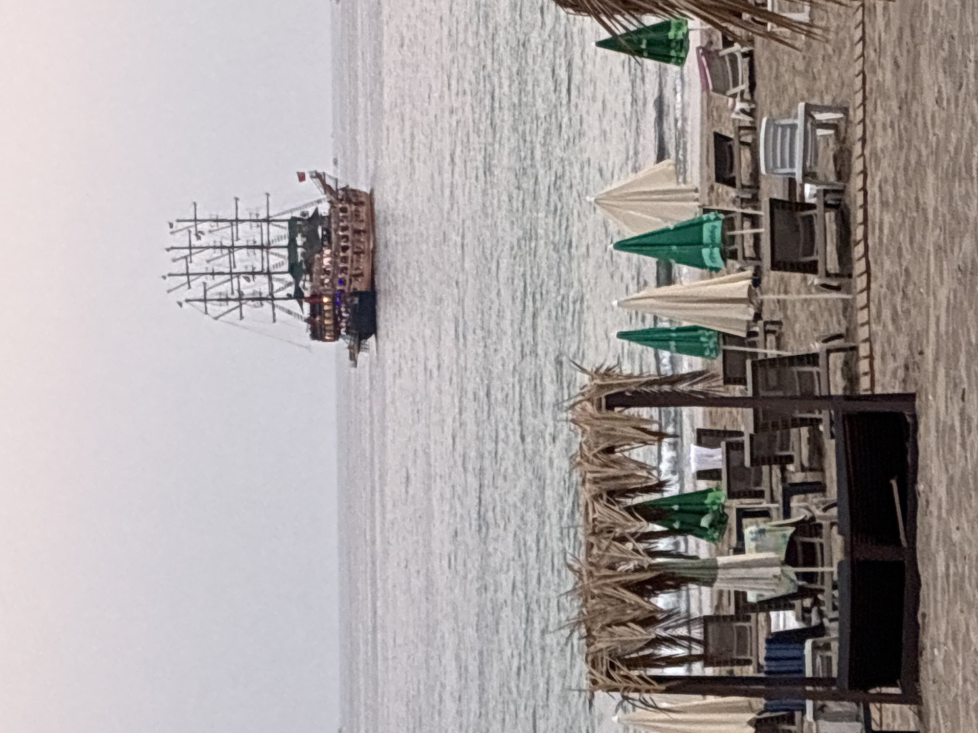 The image shows a beach in Alanya, Turkey, with a traditional pirate-style ship sailing in the distance on a calm sea under an overcast sky. In the foreground, there are several rows of empty beach chairs and umbrellas set up on the sand. A thatched-roof canopy structure with seating is also visible in the foreground. The overall scene suggests a quiet, possibly early morning or late evening setting on a beach resort.