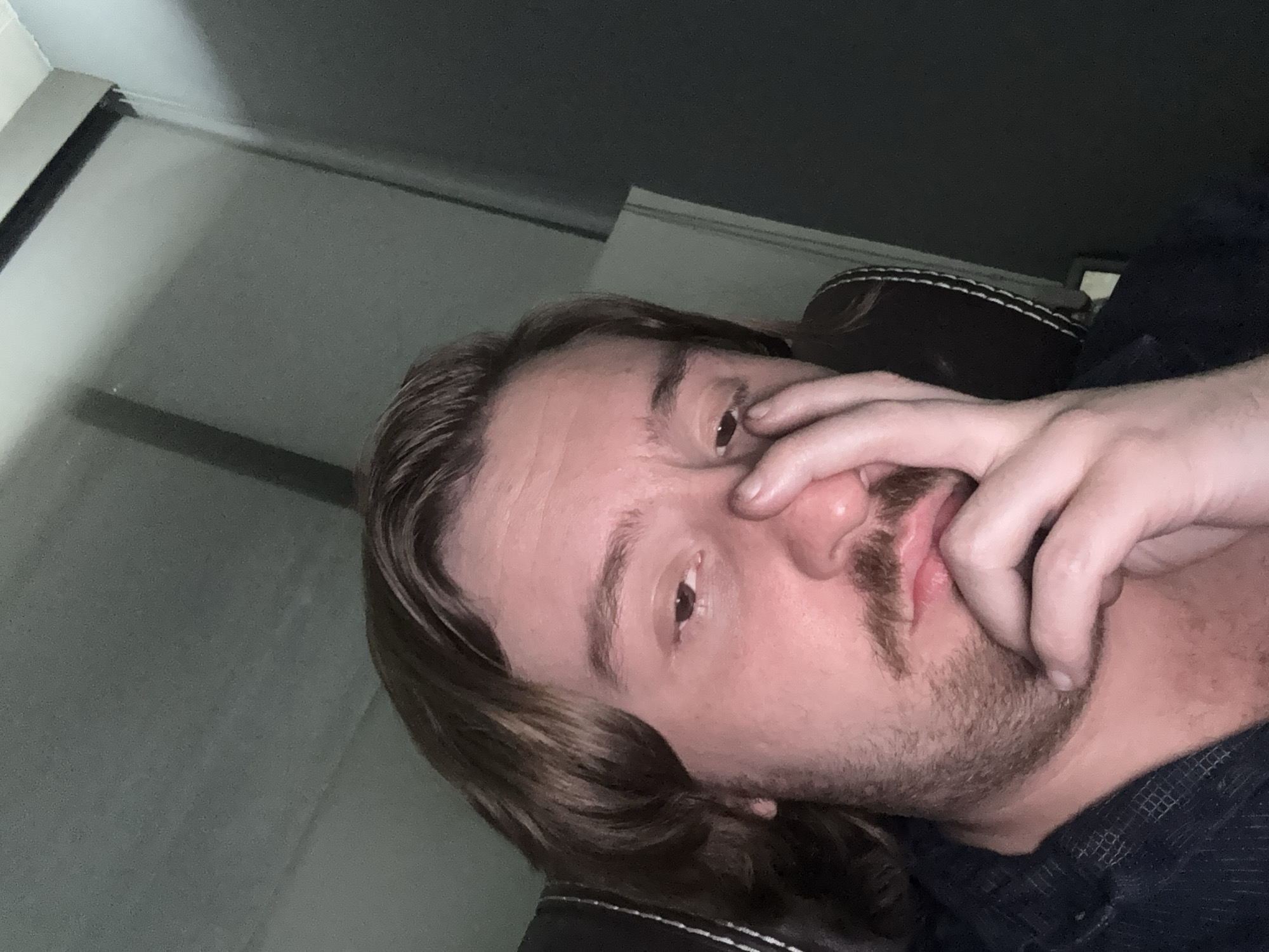 The image shows a person with long brown hair, a mustache, and a short beard. The person is looking at the camera with one eye visible; the other is partially covered by their hand, which is also touching their nose. The background is a blurry combination of a wall, a window with blinds, and what appears to be the top of a dark-colored upholstered chair or headrest. There's not enough information in the image to determine a location in Cuautitlán Izcalli, Mexico.