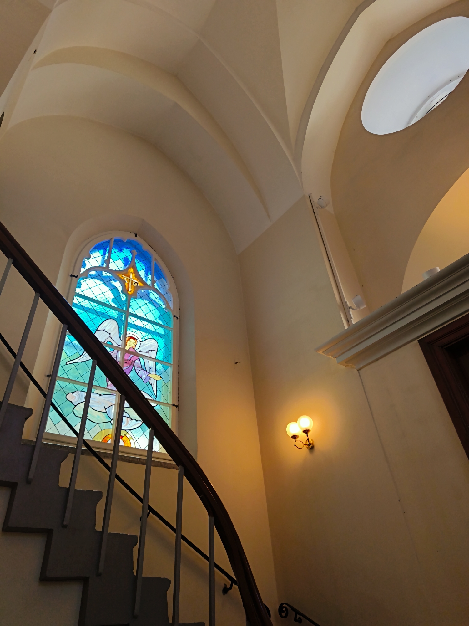 The image shows a curved staircase with a stained-glass window depicting an angel. The window is illuminated from behind, casting a colorful glow on the staircase and the surrounding cream-colored walls. The architecture features arched ceilings and a round skylight. Two wall-mounted lights with round globes are visible on the right side of the image, emitting a warm yellow light. The scene is captured from a low angle, emphasizing the upward sweep of the stairs and the vaulted ceiling. The style suggests an interior of a historical building, possibly a church or an old public building. This specific stained-glass window is located in the staircase of the Jaani kirik (St. John's Church) in Tallinn, Estonia.