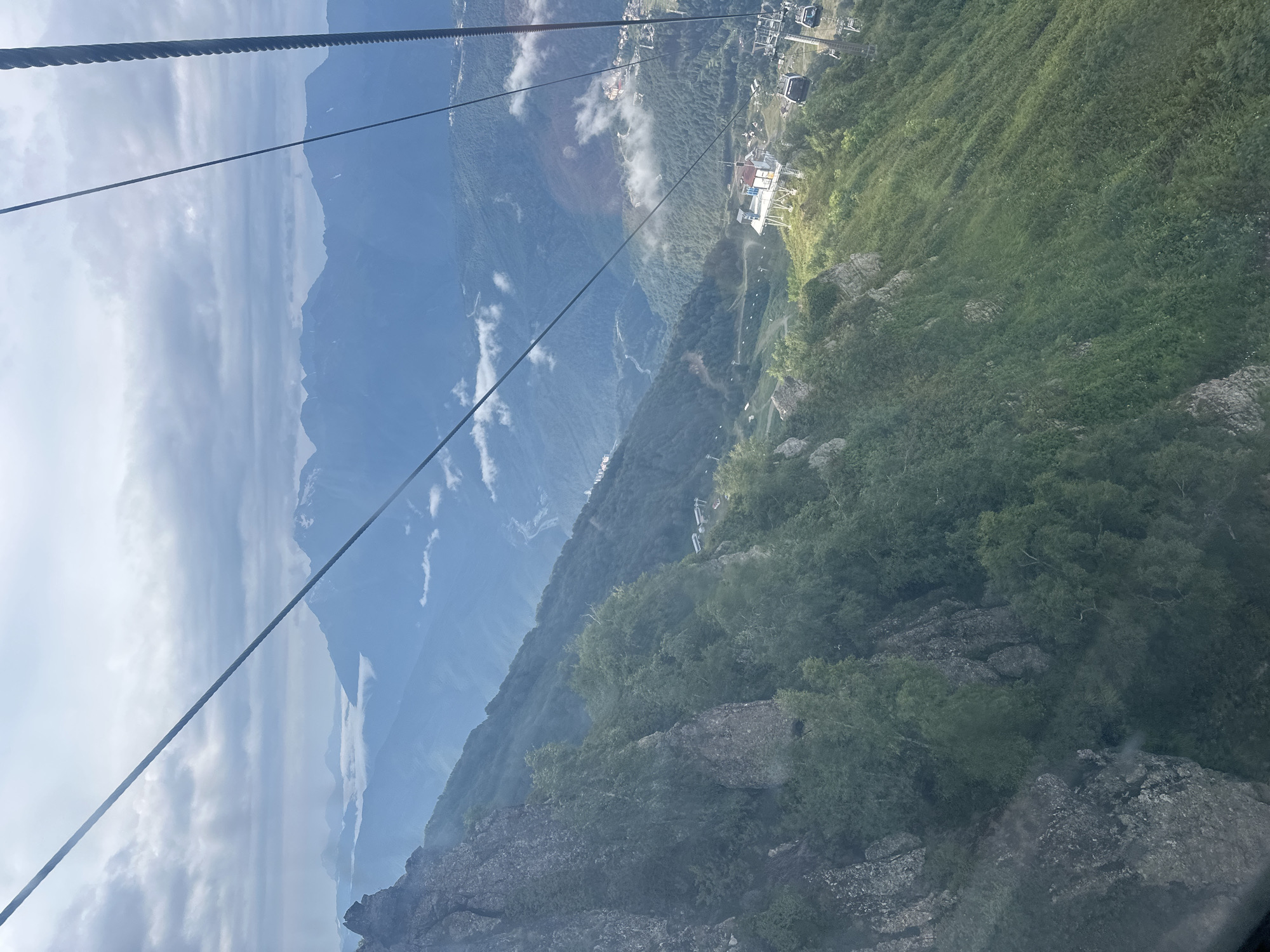 The image captures a view from a ski lift or cable car ascending a mountain in Estosadok, Russia. The foreground features a steep, rocky slope covered with dense green foliage. As the viewpoint moves upwards and away, the landscape reveals a vast expanse of rolling, forested mountains under a cloudy sky. In the mid-ground, several ski lifts and cable car cabins are visible, with one station featuring a white, multi-level structure. Farther in the distance, more mountain ranges recede into a hazy blue, with a winding river or road visible in a valley. Signs of human settlement, including buildings and infrastructure, are scattered across the mountainsides. The overall impression is a scenic mountain resort in a vast, natural setting.