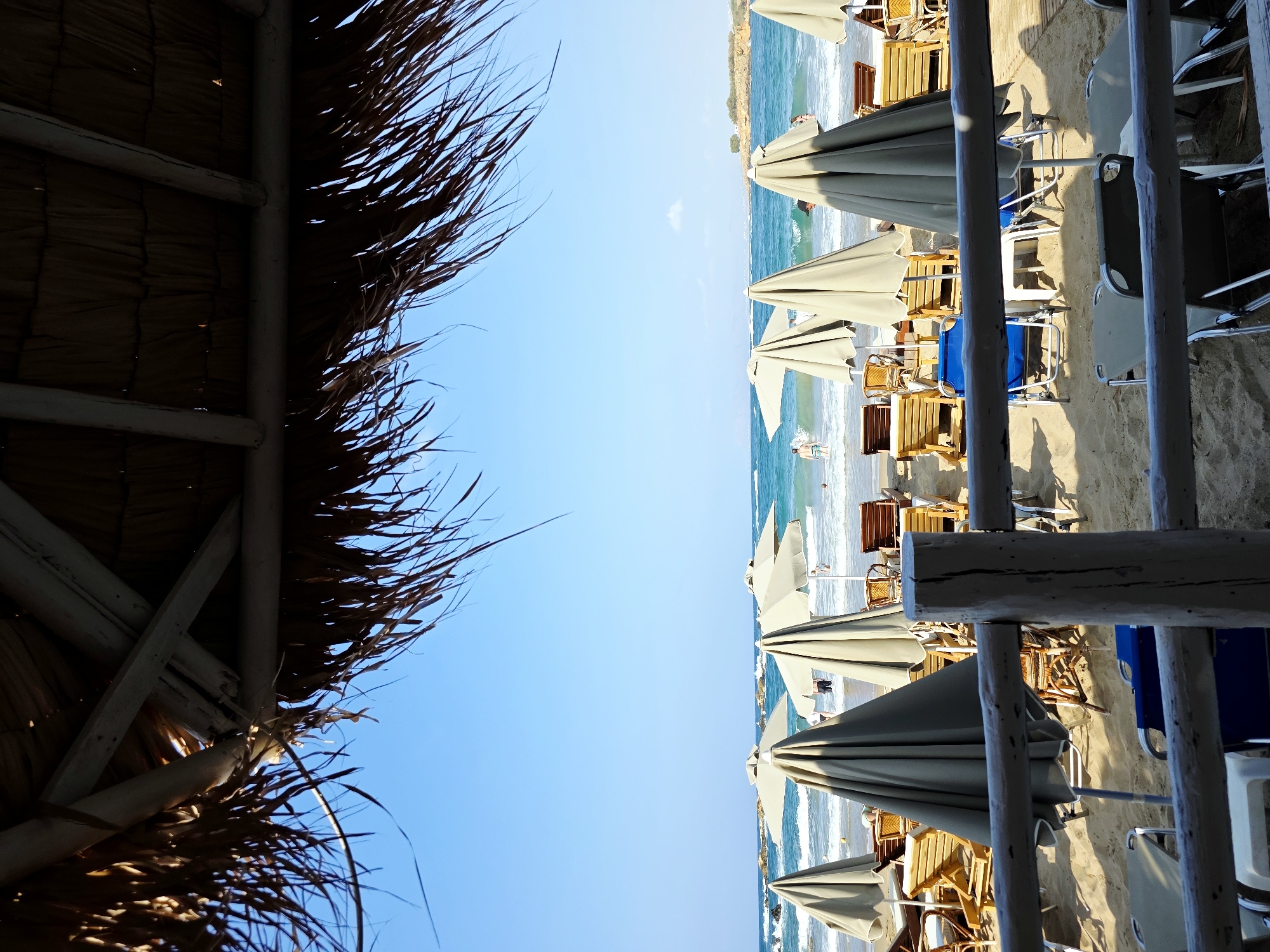 The image depicts a beach scene in Chania, Greece, viewed from beneath a thatched roof structure. The foreground is dominated by a rustic wooden railing and several beach chairs, some with blue cushions. Beyond the railing, a sandy beach is lined with numerous closed beach umbrellas and wooden lounge chairs. Further back, people are visible in the shallow waters of the turquoise sea, with gentle waves lapping the shore. The sky is a clear, bright blue, and the horizon is dotted with distant landforms. The overall atmosphere is that of a relaxing day at a seaside resort.