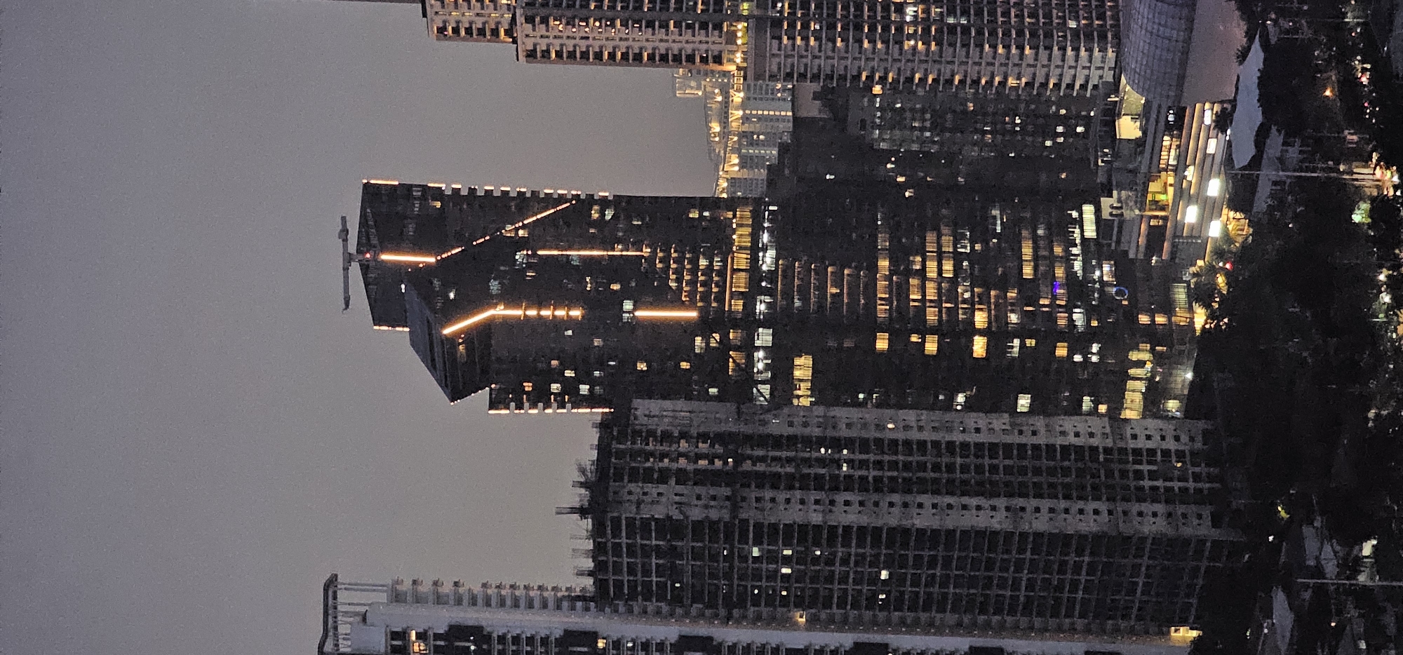 This is a high-angle, medium-shot photograph of the Makati skyline at dusk, showcasing a cluster of skyscrapers with illuminated windows. The prominent building in the center features a unique, angular architectural design with bright, vertical orange lights tracing along its facade and a horizontal structure at its apex, possibly a crane or antenna. To the left, a partially constructed building with visible rebar and floors stands starkly unfinished. To the right and in the background, other residential or office buildings exhibit a grid of warm, yellow lights. The sky is a muted grey, indicating the transition from daylight to night. The foreground is darker, with silhouettes of trees and lower structures suggesting a dense urban environment. The image captures the dynamism of urban development and nighttime illumination in Makati, Philippines.