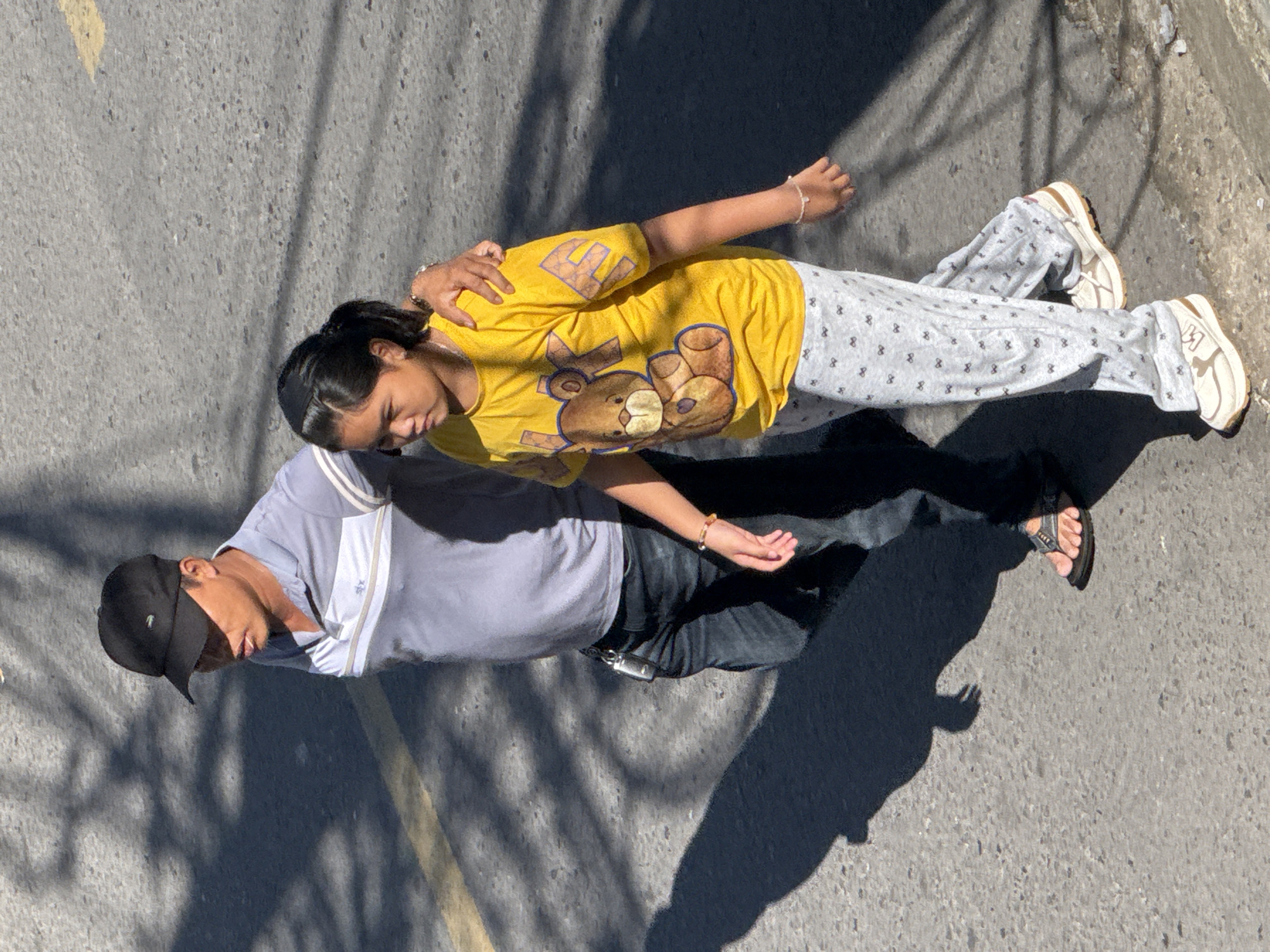 An overhead shot captures a man and a young girl walking side-by-side on a paved road in Da Lat, Vietnam. The man, wearing a black baseball cap and a light grey polo shirt with horizontal stripes on the chest, has his arm draped around the girl's shoulders. He is also wearing dark jeans and black flip-flops. The girl is dressed in a bright yellow t-shirt with a graphic of a teddy bear and the word "LOVE" printed in large purple letters. Her pants are light grey with a repeating pattern of small black bows. She wears white sneakers. The road has a faint yellow line marking a lane. The ground is textured with asphalt, and dappled sunlight casts long shadows from unseen objects, possibly trees, onto the road, creating a distinct pattern of light and dark. The lighting suggests it is daytime.