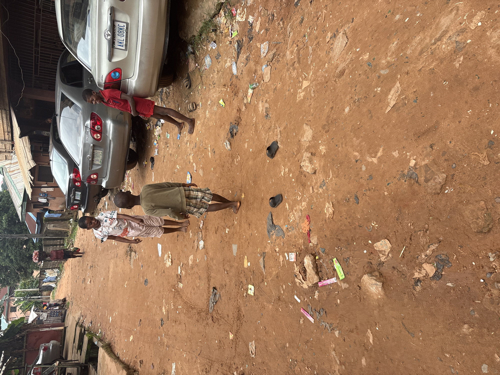 In Nkpor, Nigeria, a dirt road is lined with simple buildings and parked cars. The road surface is rough and reddish-brown, littered with various pieces of trash, including plastic bags, wrappers, and discarded footwear. In the foreground, two young boys are walking. One boy, wearing a light-colored shirt with a patterned design and light-colored shorts, faces away from the camera. Another boy, wearing a dark green long-sleeved shirt and checkered shorts, is also walking away from the camera. Further down the road, a person carries a basket of goods on their head, and a third boy, dressed in a red polo shirt and red shorts, leans against a silver Toyota Corolla. Two other cars, a dark gray Toyota Corolla and another silver car, are also visible on the road. The environment suggests a typical urban setting in a developing area, characterized by both human activity and the presence of refuse.