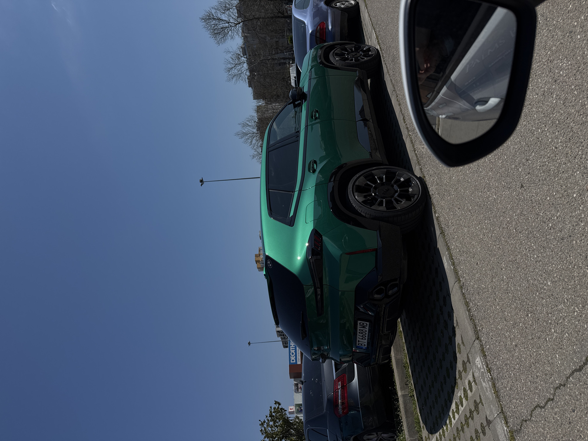 The image shows a green BMW X6 parked on the side of a road, next to other parked cars. The car has a license plate with the registration "BT 6688 MB". The side mirror of another vehicle is visible in the right foreground. In the background, there is a Decathlon store, suggesting the location is likely in an area with retail businesses. Given the license plate registration, the location is likely Burgas, Bulgaria. The sky is blue and clear, indicating it is daytime. There are no visible people.