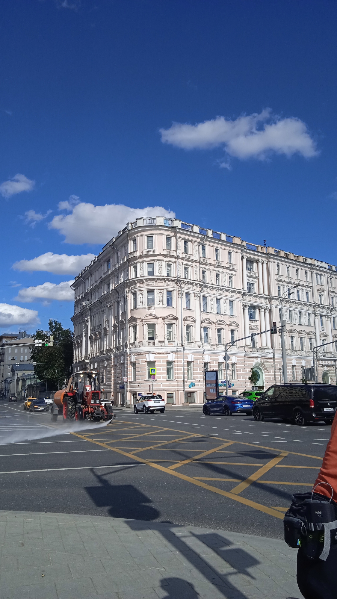 A street scene in Moscow, Russia, depicts a large, ornate, light pink building with many windows and decorative elements. In the foreground, a red tractor with a water sprayer is cleaning the road, creating a mist. Several cars are visible on the road, including a white SUV, a blue sports car, and a black van. The sky is blue with scattered white clouds. The scene appears to be a street intersection with yellow box markings on the road.