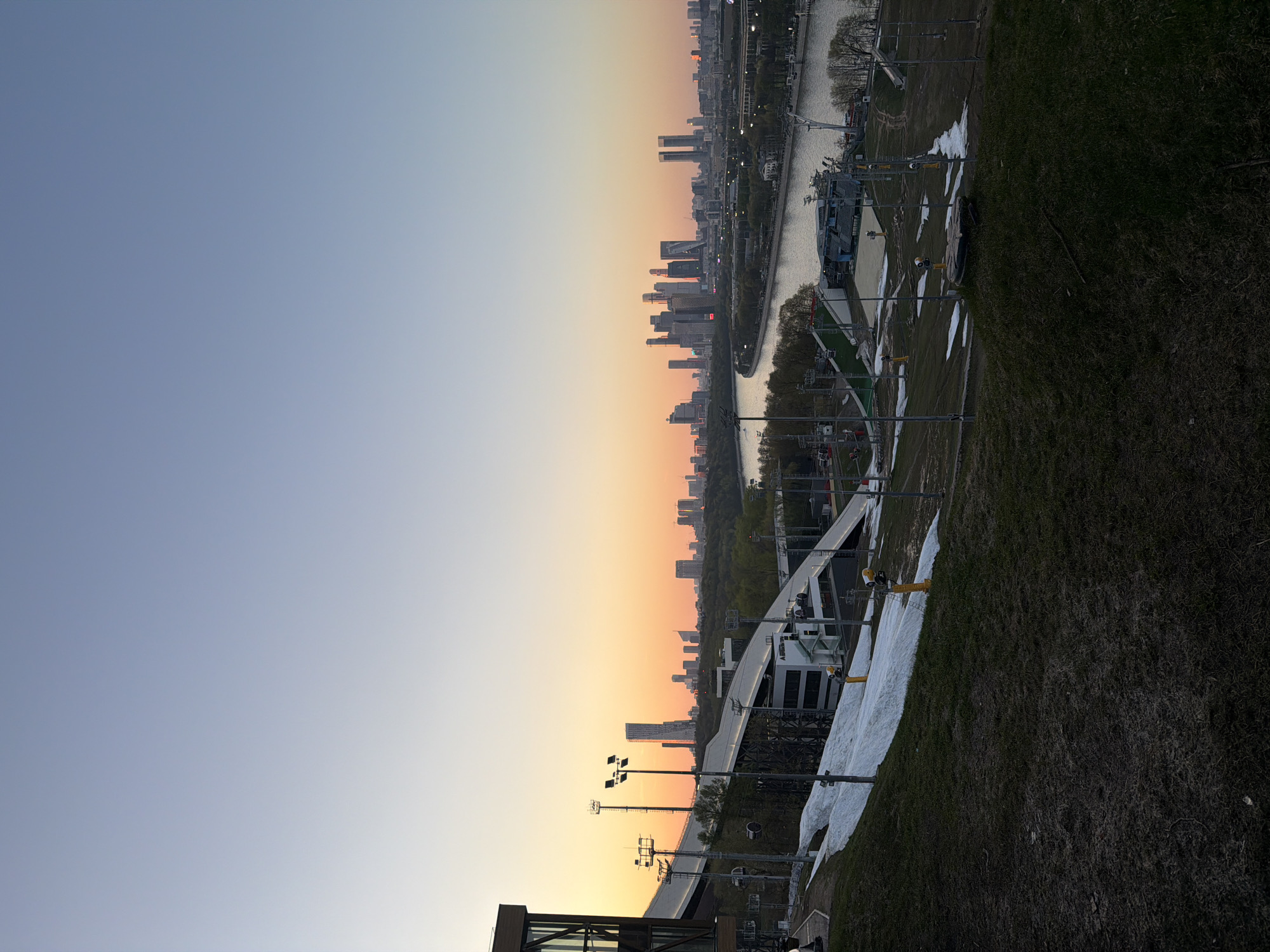 The image shows an elevated view of Moscow, Russia, at sunset. The sky transitions from blue to orange, illuminating the city skyline. A river winds through the urban landscape, with buildings lining its banks. In the foreground, there's a grassy area, possibly a park, with snow patches and light poles. The architecture includes a mix of residential and commercial buildings. A ski resort with snow is visible.