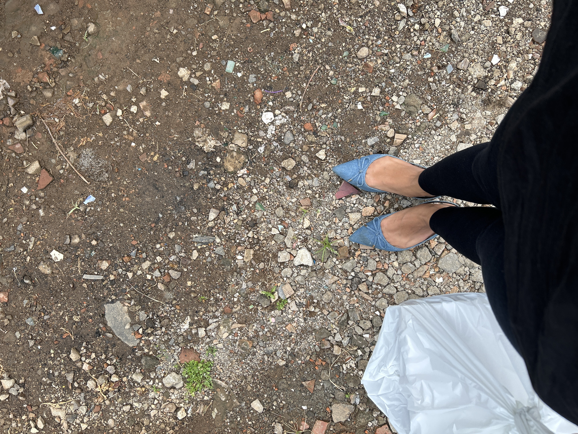 The image shows a person's legs and feet, wearing blue denim pointed-toe flats and black leggings. The person is standing on a surface composed of dirt, small stones, and fragments of brick and other debris. Some small green plants are growing through the dirt in patches. A large white plastic bag is visible in the lower right corner of the frame. The scene appears to be outdoors, in an unpaved area, possibly a construction site or a neglected urban space. The lighting suggests it could be daytime, with overcast skies. There are no discernible signs or text in the image. The location context provided is East Jakarta, Indonesia, which aligns with the appearance of the ground cover.