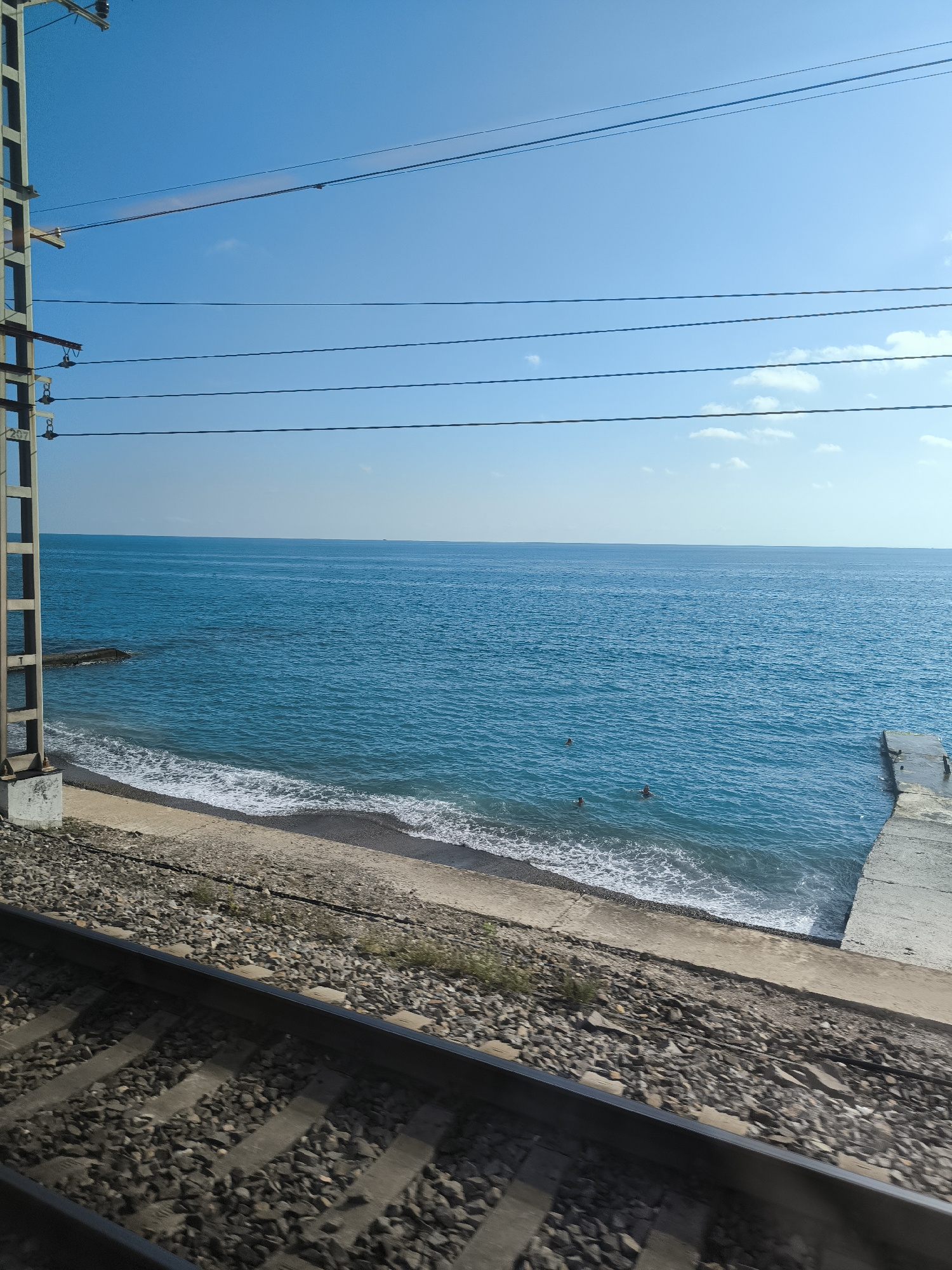 This is a landscape photograph captured from a low angle, possibly from a moving train. In the foreground, train tracks are visible, composed of dark gravel and concrete sleepers, with the metal rails glinting. To the left of the tracks, a tall, grey utility pole stands, with visible wires stretching horizontally across the frame.
Beyond the tracks and pole, a beach transitions into the sea. The beach itself appears to be a mix of sand and small dark pebbles, with a white foamy shoreline where waves are breaking. The sea is a vibrant turquoise blue, stretching out to the horizon. A small concrete pier extends into the water on the right side of the frame. In the water, three indistinct figures can be seen swimming. The sky above is a clear, bright blue, with a few wispy white clouds. The overall scene suggests a coastal location with active railway infrastructure adjacent to the beach.