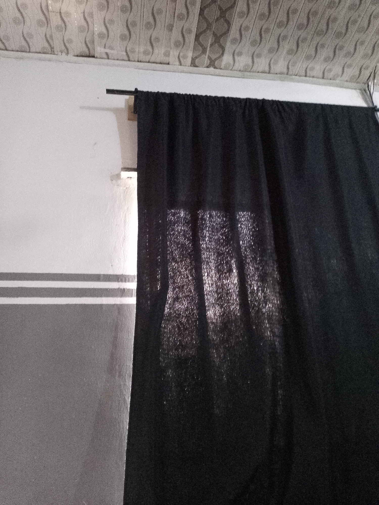 The image shows a black curtain hanging from a curtain rod against a white and grey wall. The ceiling above has a patterned design. The curtain is partially translucent, allowing light to filter through and create a shimmering effect. There are no people or animals visible in the image. The location is not specified in the image.