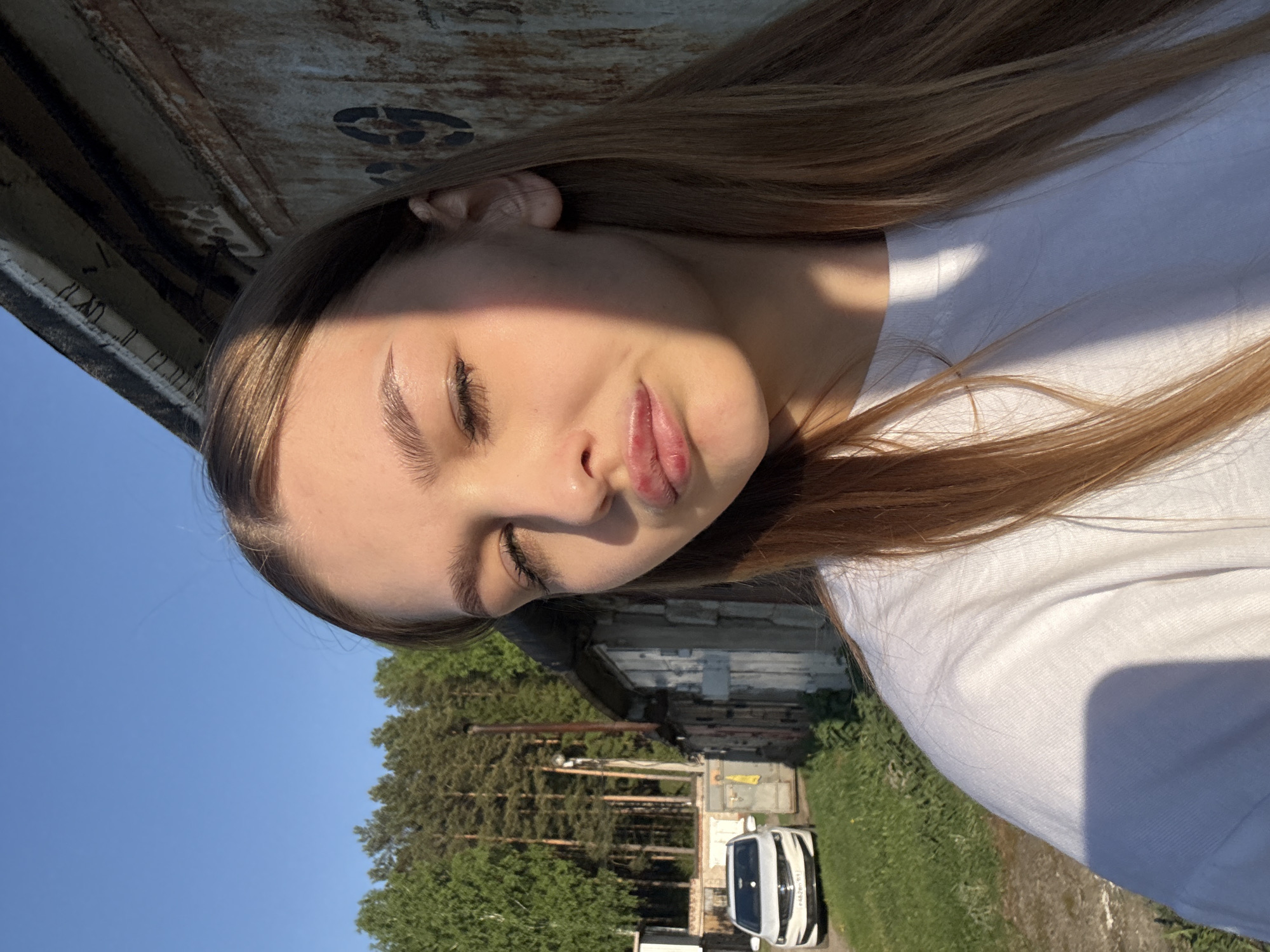 A young woman lies in an outdoor location in Chaikovsky, Russia. Her head rests against what appears to be a large, metal structure, possibly a container or part of a building. A white car is visible in the background, parked on a road lined with green vegetation. Trees are also present in the background. The sky is blue.
