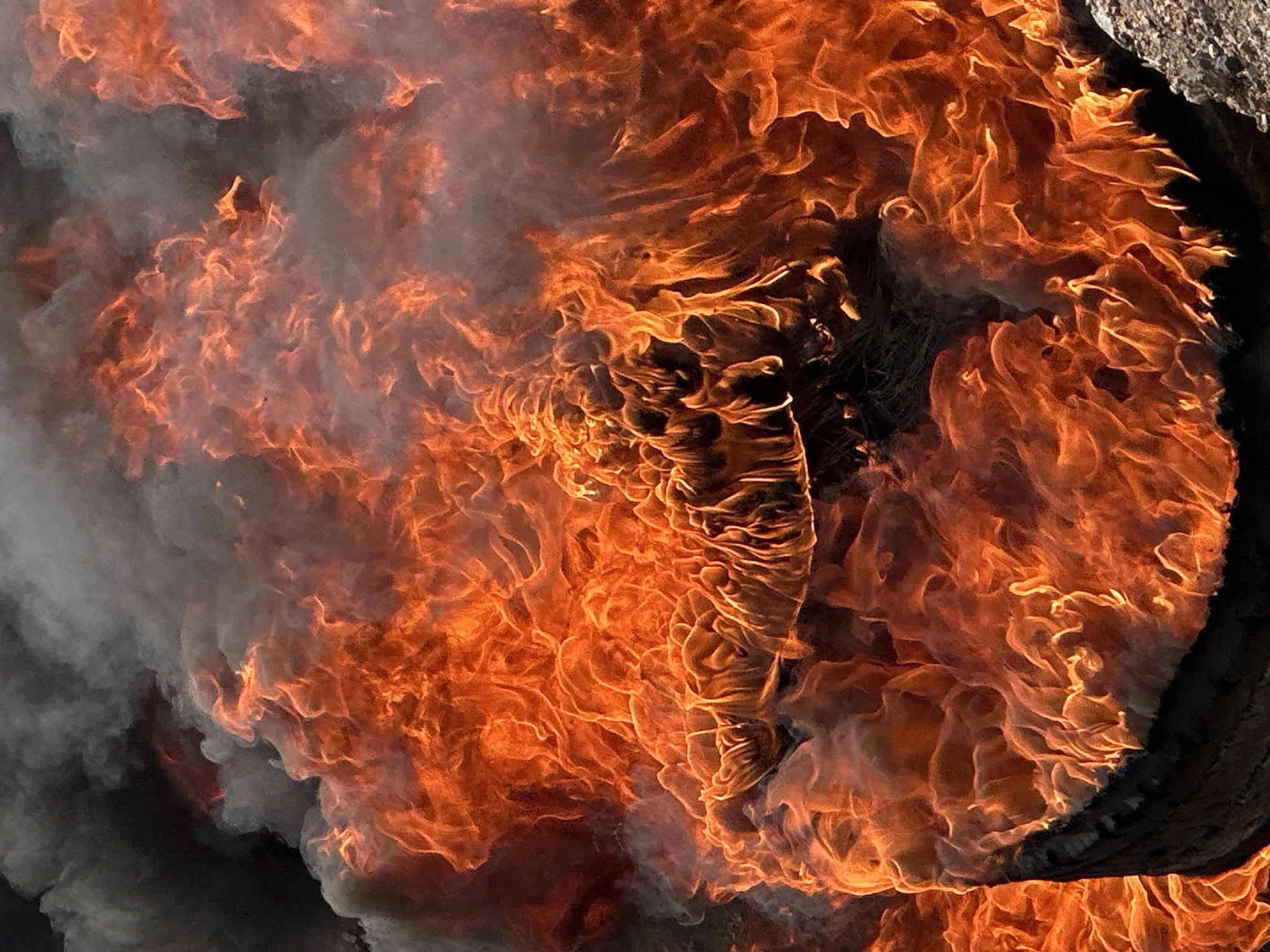 The image displays a close-up view of a tire engulfed in intense flames and smoke. The fire is the dominant element, characterized by vibrant oranges, yellows, and reds, with fluid, swirling patterns. Dark gray smoke billows and intermingles with the flames, obscuring parts of the scene and adding a sense of chaos. The tire itself is partially visible at the bottom and left edges, appearing charred and distorted by the heat. The texture of the burning material is rough and irregular. The background is a dark, indistinct mass, suggesting a smoky, uncontrolled fire. There are no people or specific landmarks visible that would indicate the location in Gura Top, Nigeria. The scene is purely focused on the destructive power of the fire consuming the tire.
