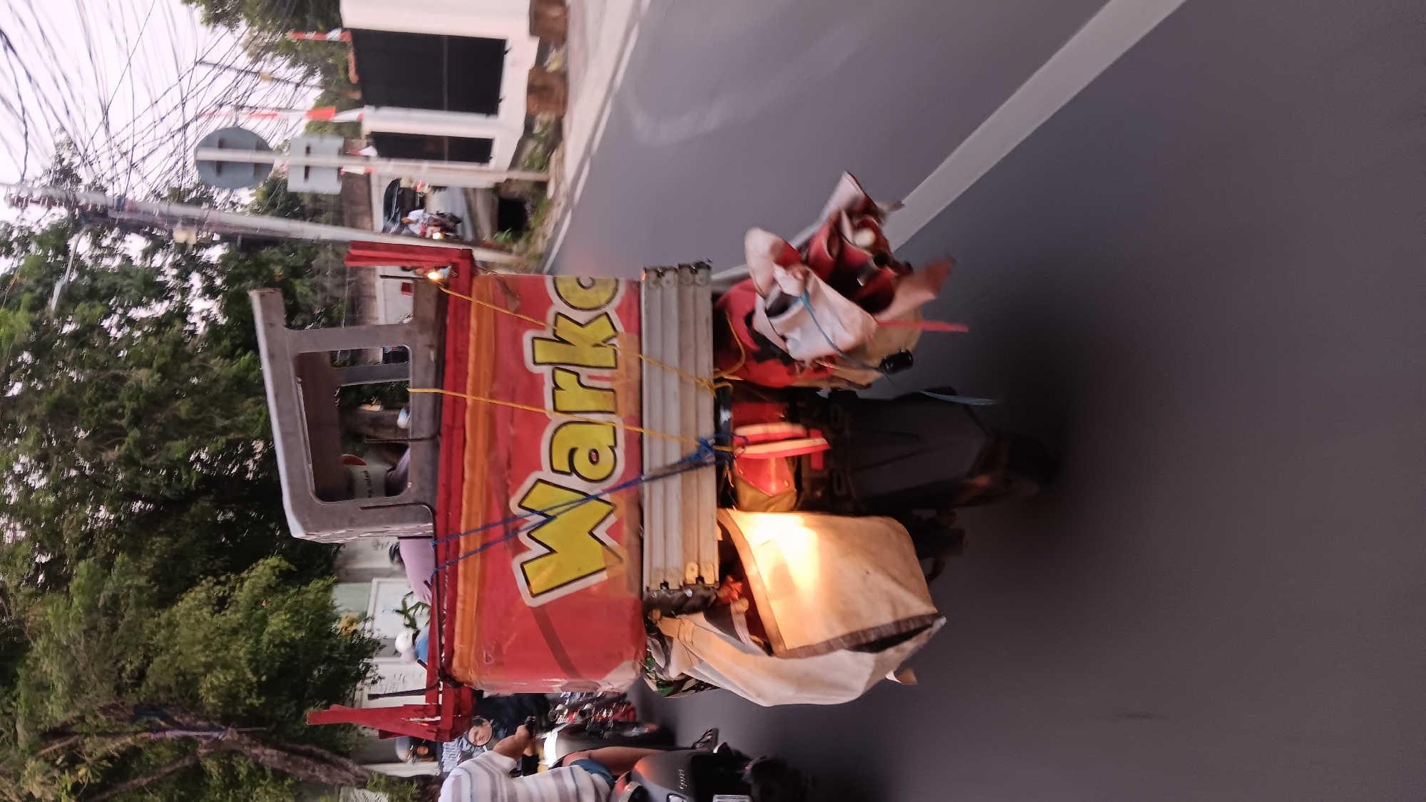 A motorcycle with a large red cargo box on its rear is driving on a paved road in South Jakarta, Indonesia. The cargo box has "Warkop" written on it in yellow letters outlined in black. The box is loaded with various items, including bags, clothing, and a metal frame on top. There are also several other motorcycles and pedestrians on the road. The area appears to be a residential or commercial district with trees and buildings lining the street. The sky is overcast, and the lighting suggests it is either early morning or late afternoon.