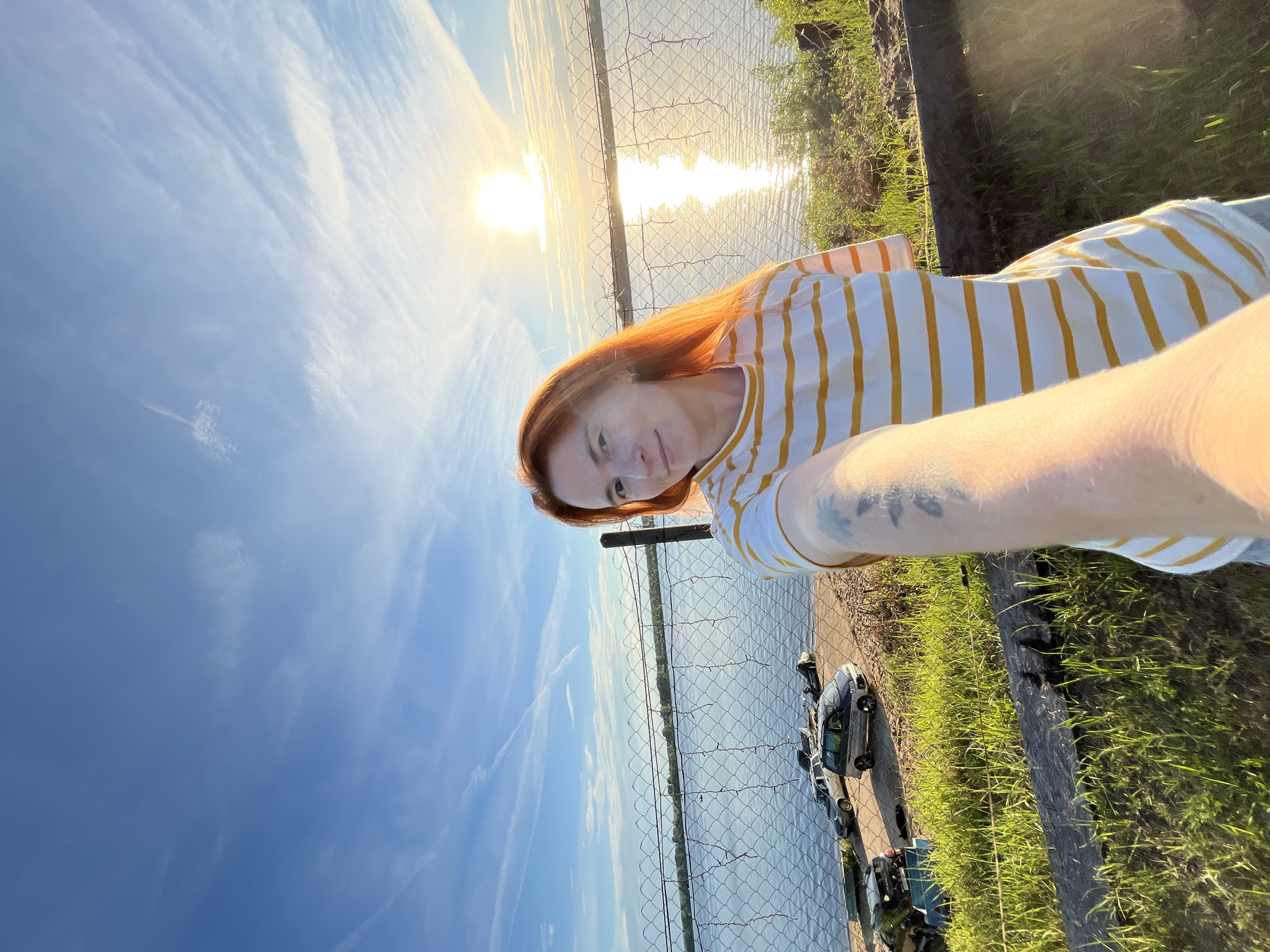 A woman with red hair, freckles, and a striped t-shirt takes a selfie outdoors. She is smiling slightly and her arm is extended to capture the photo. Behind her, a chain-link fence separates the foreground from a body of water, likely a river, that reflects the bright sunlight. The sun is low in the sky, casting a warm glow and creating a shimmering effect on the water, suggesting it is late afternoon or early evening. The sky is a vibrant blue with wispy white clouds. To the right of the woman and the fence, there is green grass and a wooden post. A few small vehicles are parked on the grassy area next to the water. The overall mood of the image is relaxed and serene. The image context indicates the location is Chaikovsky, Russia. There is no visible text in the image.