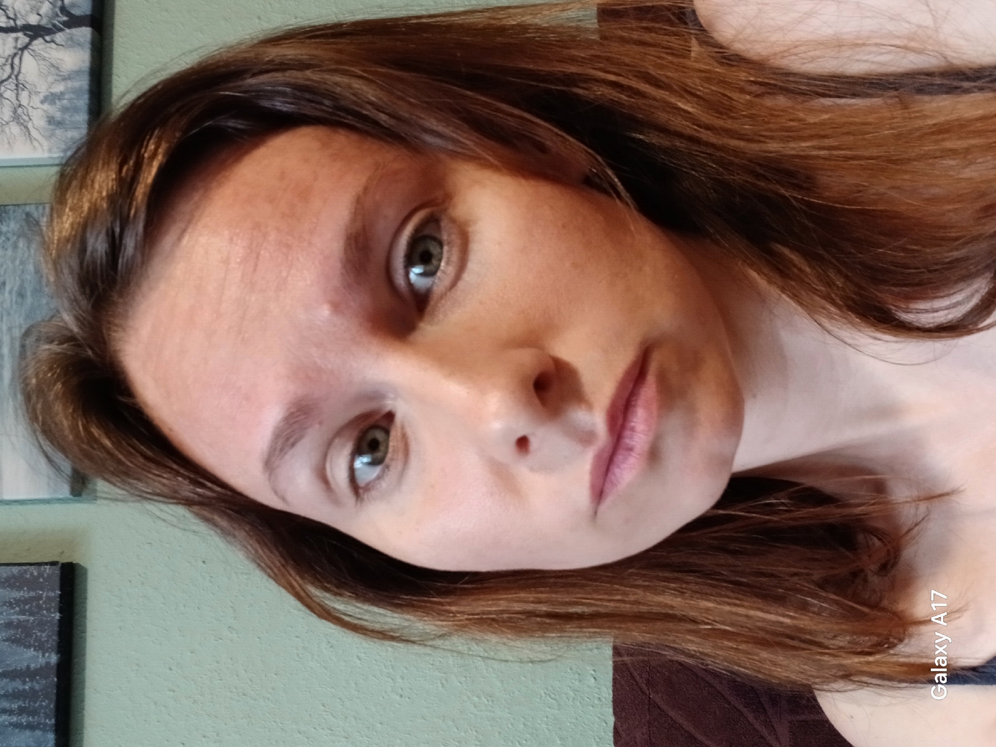 A woman with light skin and light brown hair looks directly at the camera. Her eyes are light green. She has light pink lips. The background appears to be a light green wall with a framed picture. The text "Galaxy A17" is visible in the bottom right corner of the image. There is no indication this image was taken in Yekaterinburg, Russia.