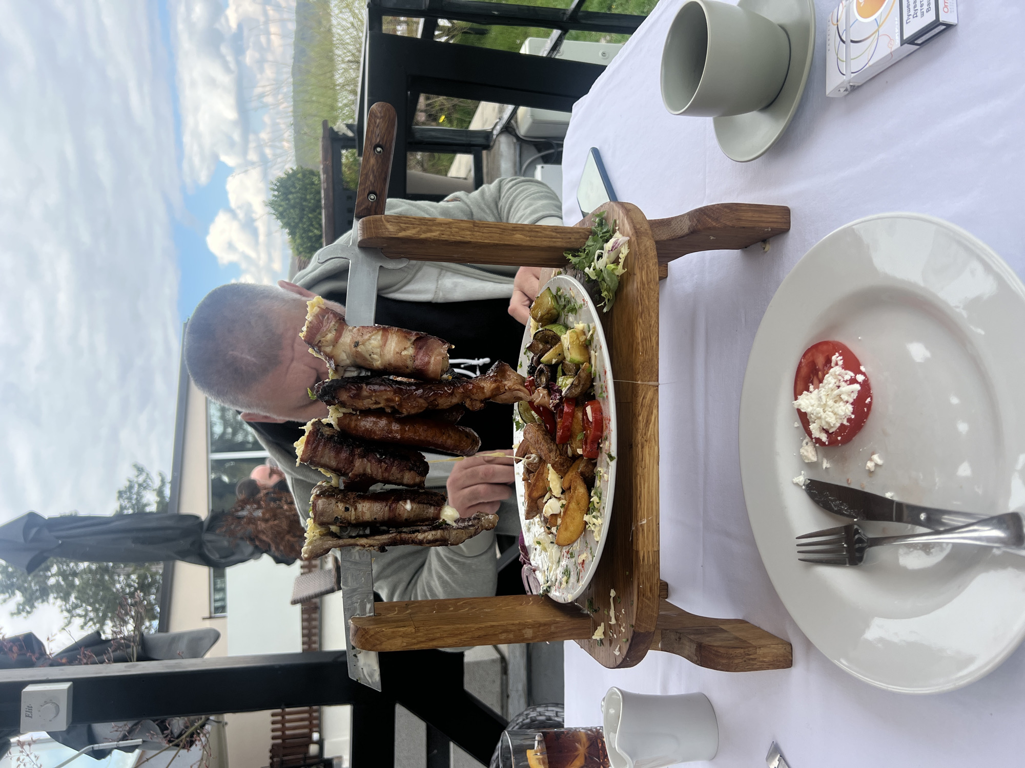 A man is eating a large meal presented on a wooden stand at a table in what appears to be an outdoor restaurant. The meal consists of multiple skewers of meat wrapped in bacon, served above a plate of assorted vegetables. To the right of the wooden stand are a plate with a sliced tomato topped with crumbled cheese, a fork and knife, a grey cup and saucer, and a white cigarette pack. The table is covered with a white tablecloth. The man is wearing a grey jacket. In the background, trees, buildings, and the sky are visible. The location is likely in the Smederevska Palanka Municipality, Serbia.