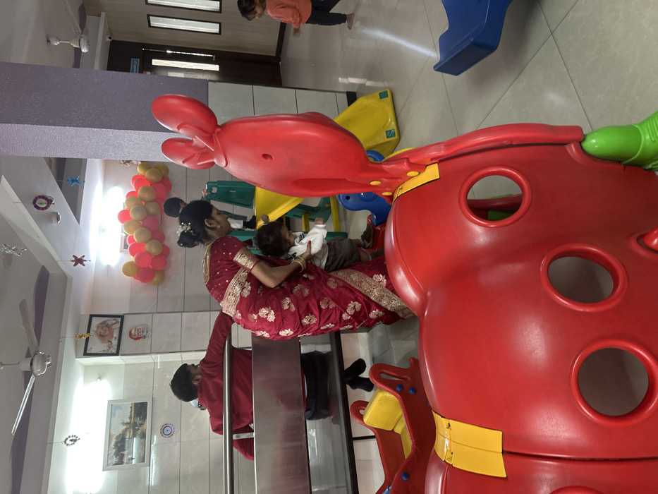 A woman in a maroon saree holds a baby on her lap near a stainless steel railing. A man in a red shirt stands nearby. A red and yellow playground structure dominates the foreground. A yellow and blue slide and a person standing are visible in the background. Wall decorations, a ceiling fan, and a balloon arrangement are also present. The image is rotated counterclockwise.