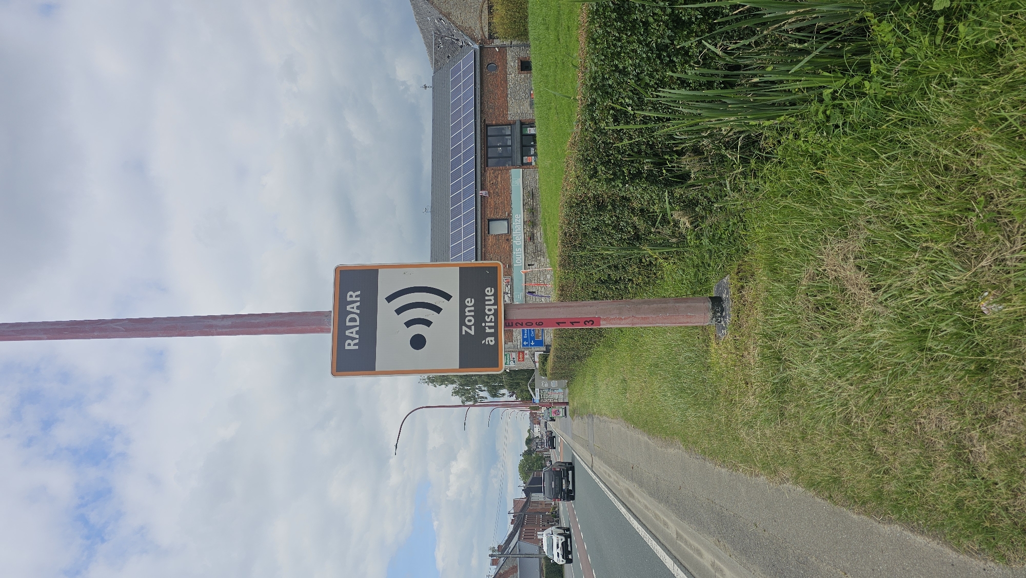 A radar speed camera sign is mounted on a red pole by the side of a road in Boussu-lez-Walcourt, Belgium. The sign is rectangular with an orange border and a white background. It displays the word "RADAR" in black letters at the top, followed by a black radar symbol and the text "Zone à risque" (Risk zone) in black. A white arrow pointing left with "Deneu" and an arrow pointing up with "Boussu" are visible on a smaller blue sign below the radar sign. Red markings, E206 and 113, are visible vertically on the pole. The road is paved and has a white line marking. Several vehicles, including a white car and a dark SUV, are driving on the road. A building with a brick facade and solar panels on the roof is situated behind a green hedge. The environment is characterized by green grass and vegetation on the right side of the road. The sky is cloudy with patches of blue.