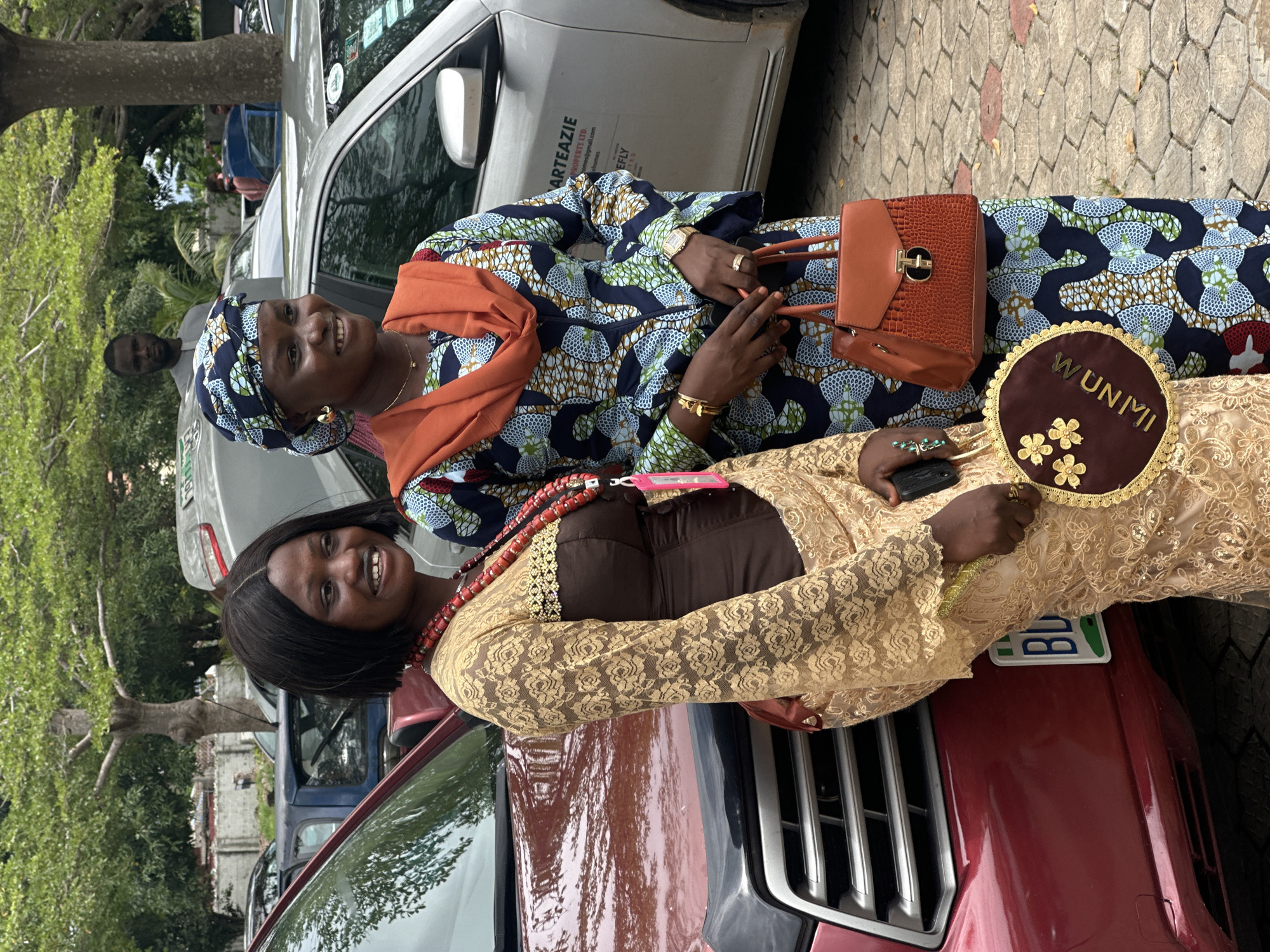 In the city of Itoga, Nigeria, two women are standing next to a red vehicle. The woman on the left is wearing a gold lace overlay over a brown dress, a red beaded necklace, and a brown handbag. She is holding a decorative circular fan with gold floral embellishments and the word "WUNMI" written in gold lettering. The woman on the right is wearing a blue, white, and green patterned dress with an orange headscarf and shawl. She is holding an orange textured handbag. A man is visible in the background, standing behind some cars. The environment appears to be an outdoor parking area, with trees and paved ground.